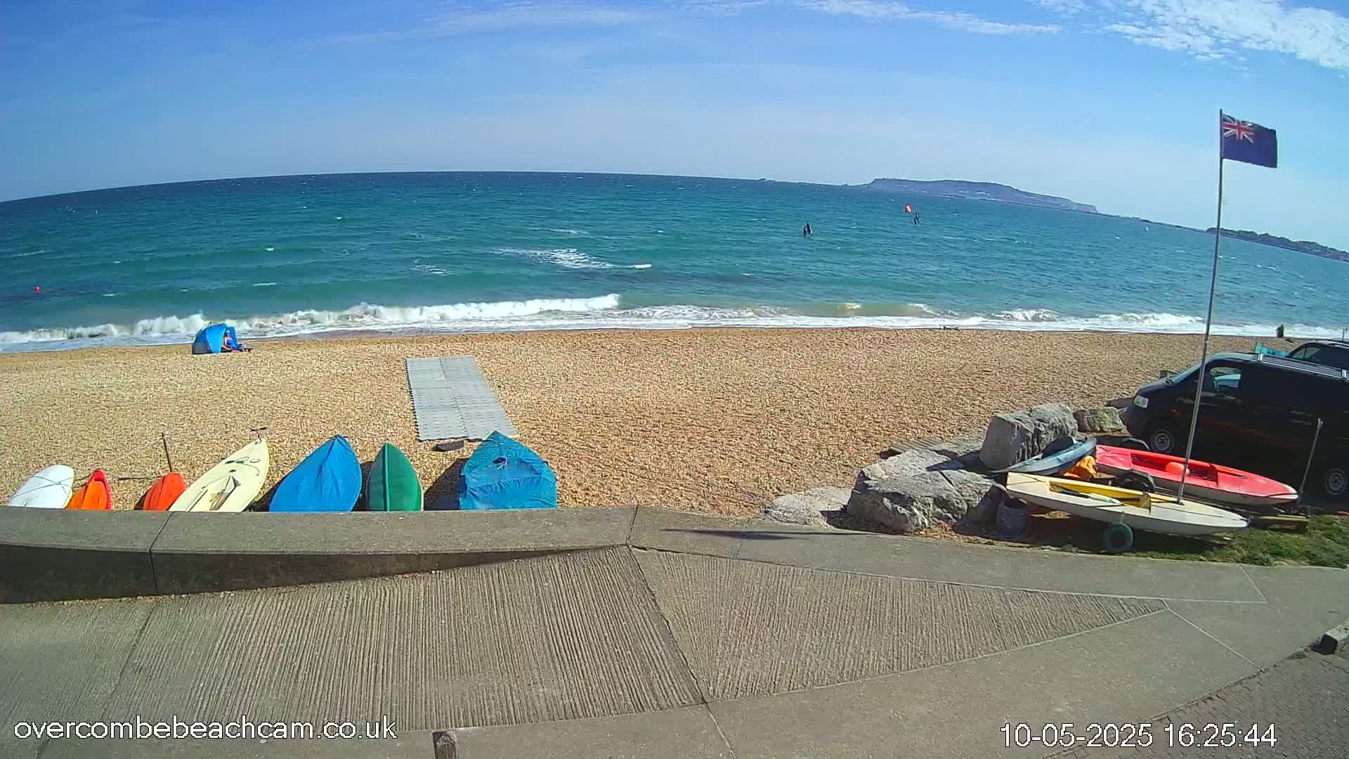 A sandy beach with several small boats resting on the shore, a blue tent, and a parked van, overlooks a calm sea with a few small waves under a sunny sky.