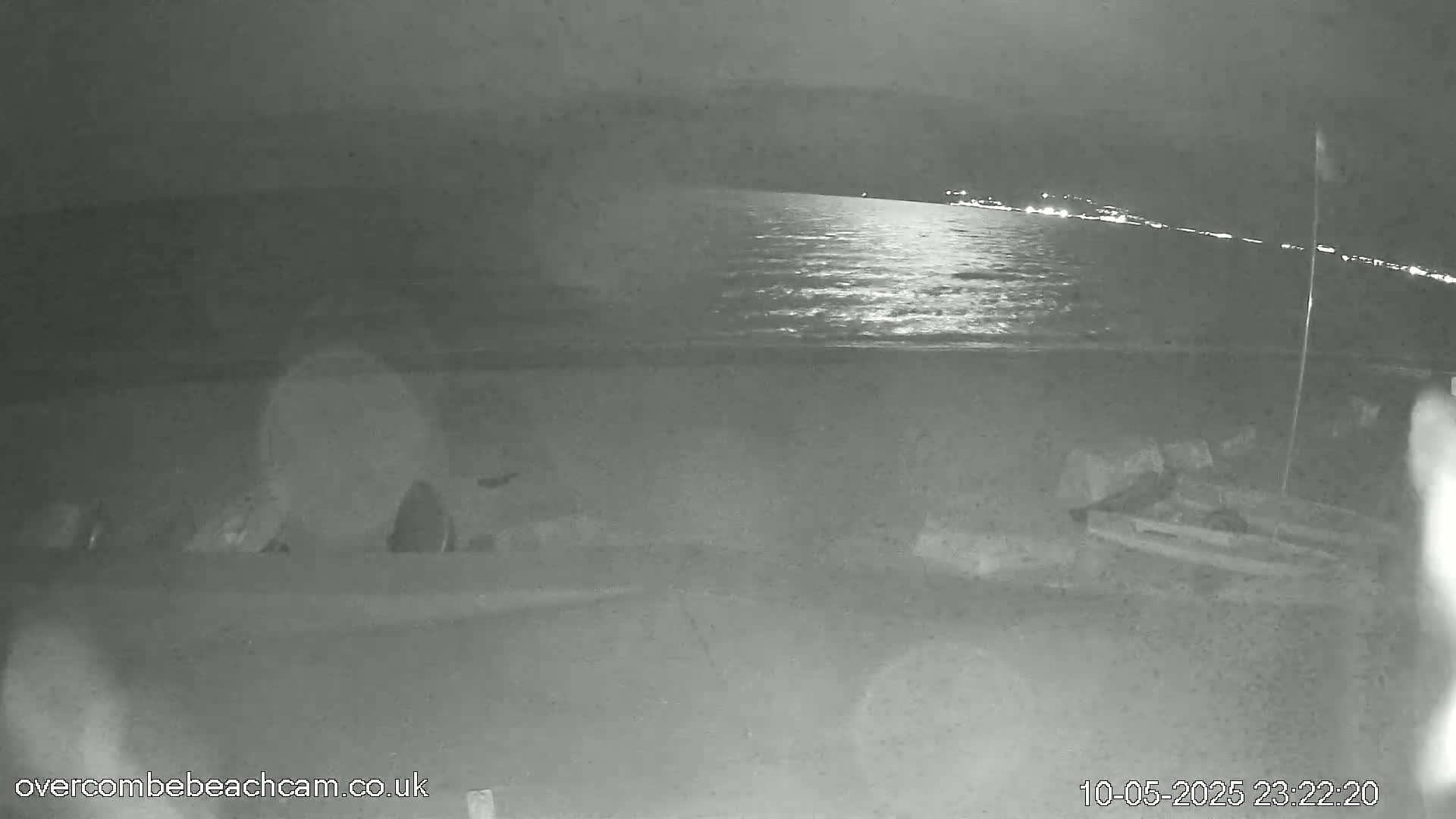 A nighttime, low-light image shows a dark beach with a calm sea reflecting light, and some indistinct objects on the sand.