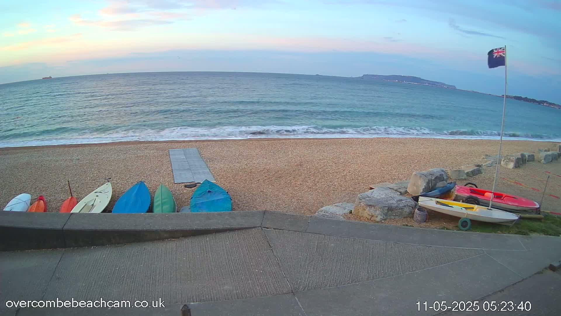Several small boats sit on a pebble beach next to a calm sea under a mostly clear sky at dawn.