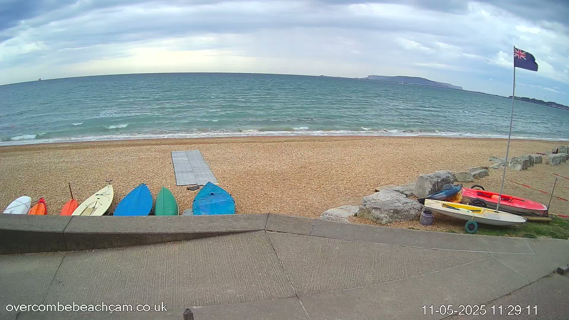 Several small boats are lined up on a pebble beach next to a calm sea under a cloudy sky.