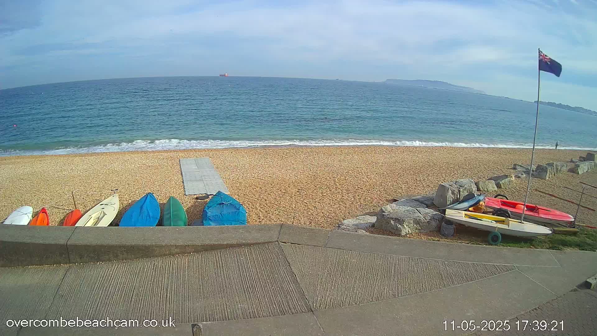 Several small boats are lined up on a pebble beach next to a calm, blue ocean under a partly sunny sky.