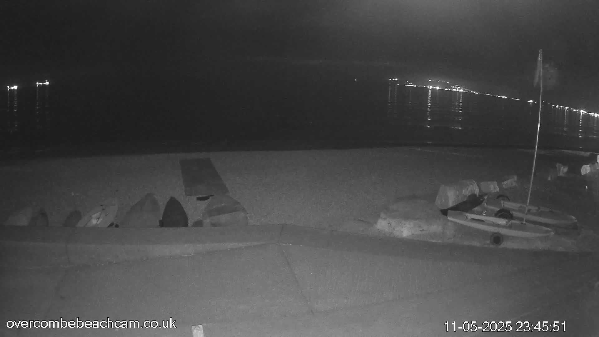 A nighttime, low-light beach scene shows several objects lying on the sand near a low wall, with a small boat and a flagpole near the water's edge, and distant lights visible across the water.