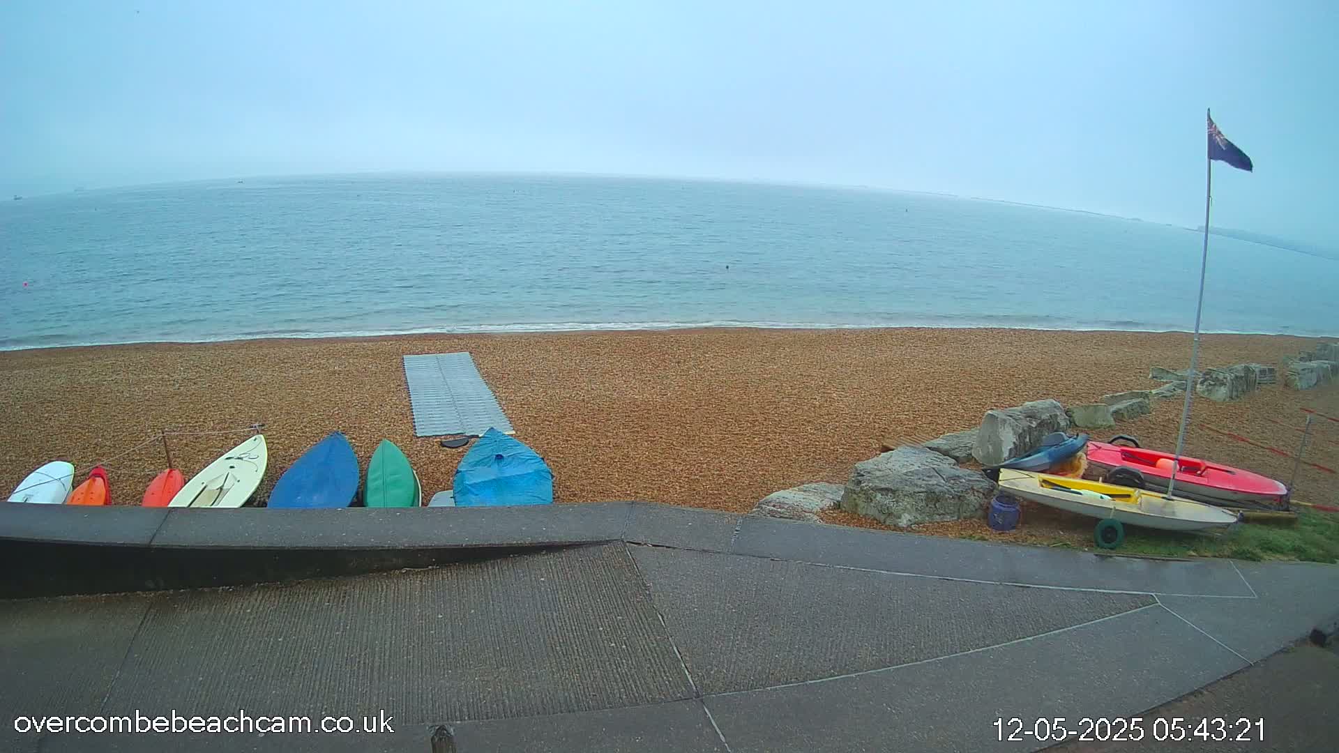 Several small boats are lined up on a pebble beach next to a calm, foggy sea.