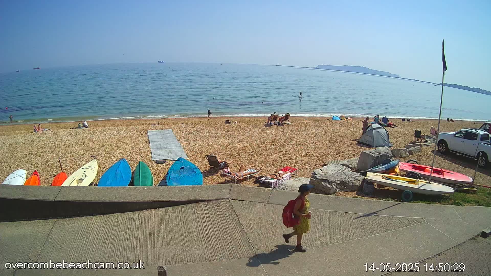A sunny beach scene shows people sunbathing, swimming, and relaxing near colorful kayaks and sailboats, with a calm ocean in the background.