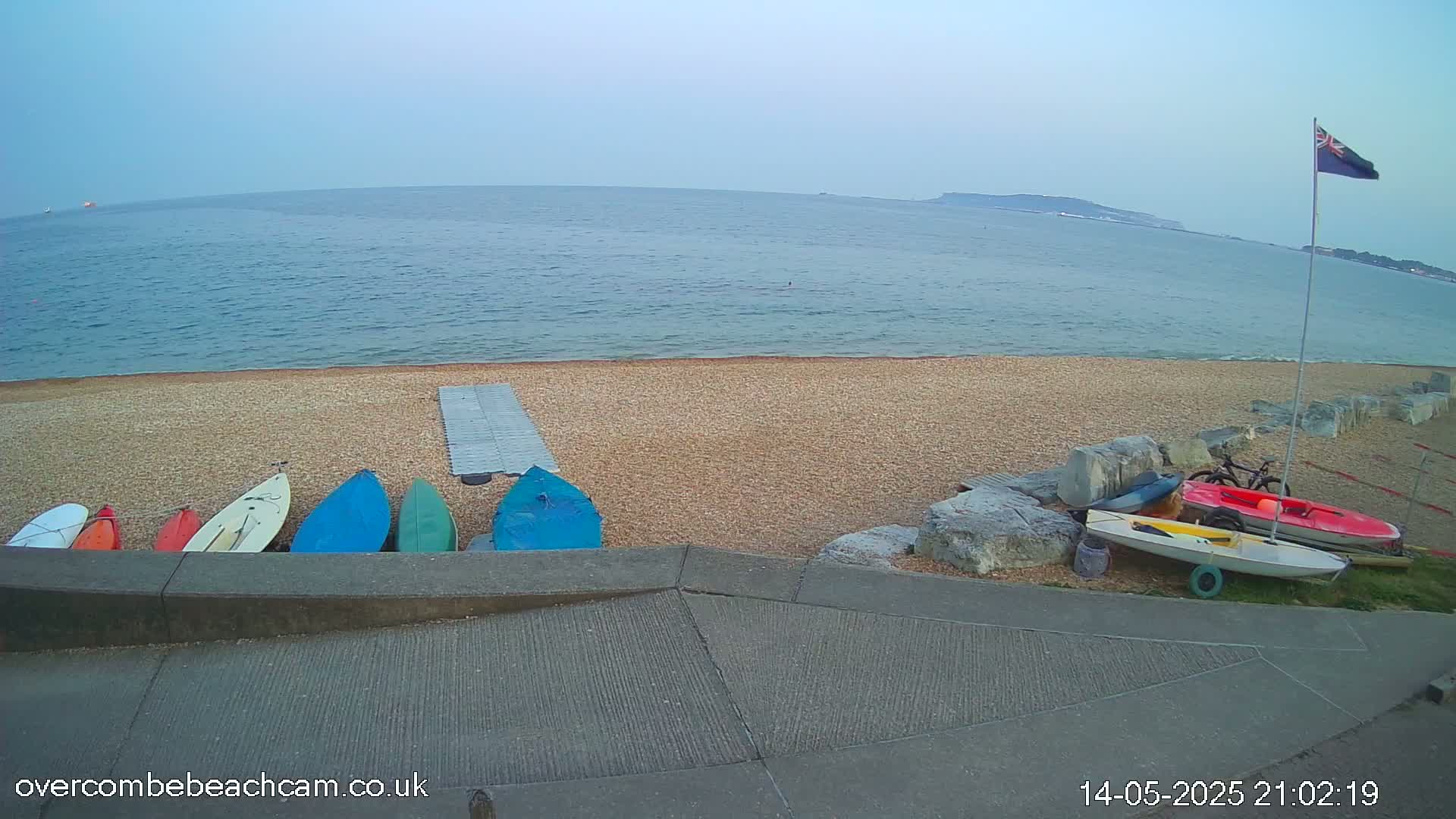 Several small boats and kayaks are resting on a pebble beach next to calm, blue water under a twilight sky.