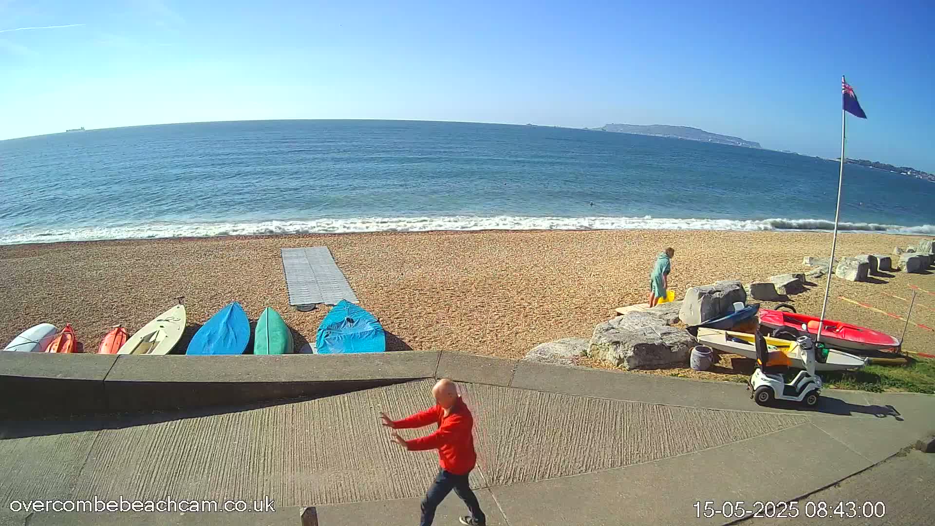 A man in a red jacket stands on a paved path overlooking a pebble beach with several kayaks and a mobility scooter, under sunny skies and calm ocean waves.