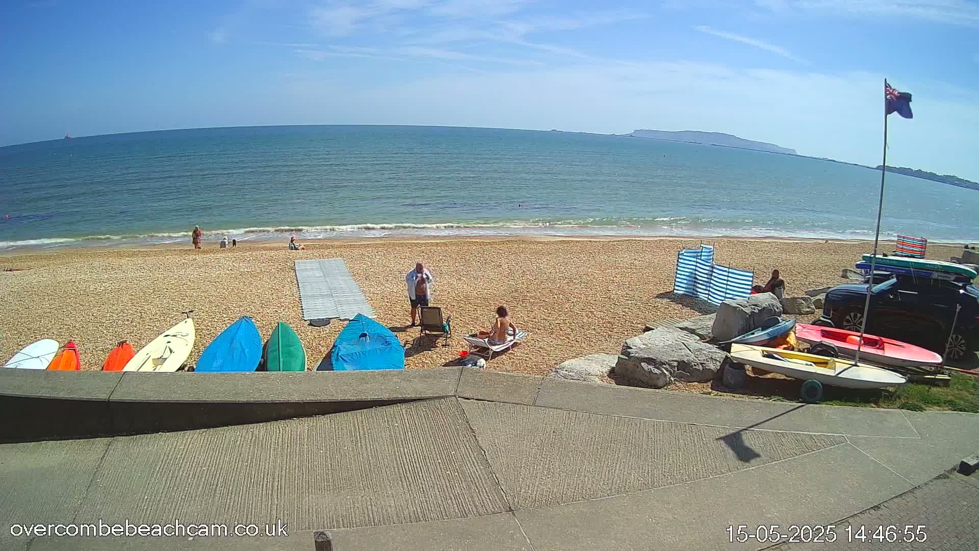 A sunny beach scene shows several people relaxing near colorful kayaks and sailboats on a sandy beach, with a calm ocean extending to the horizon.