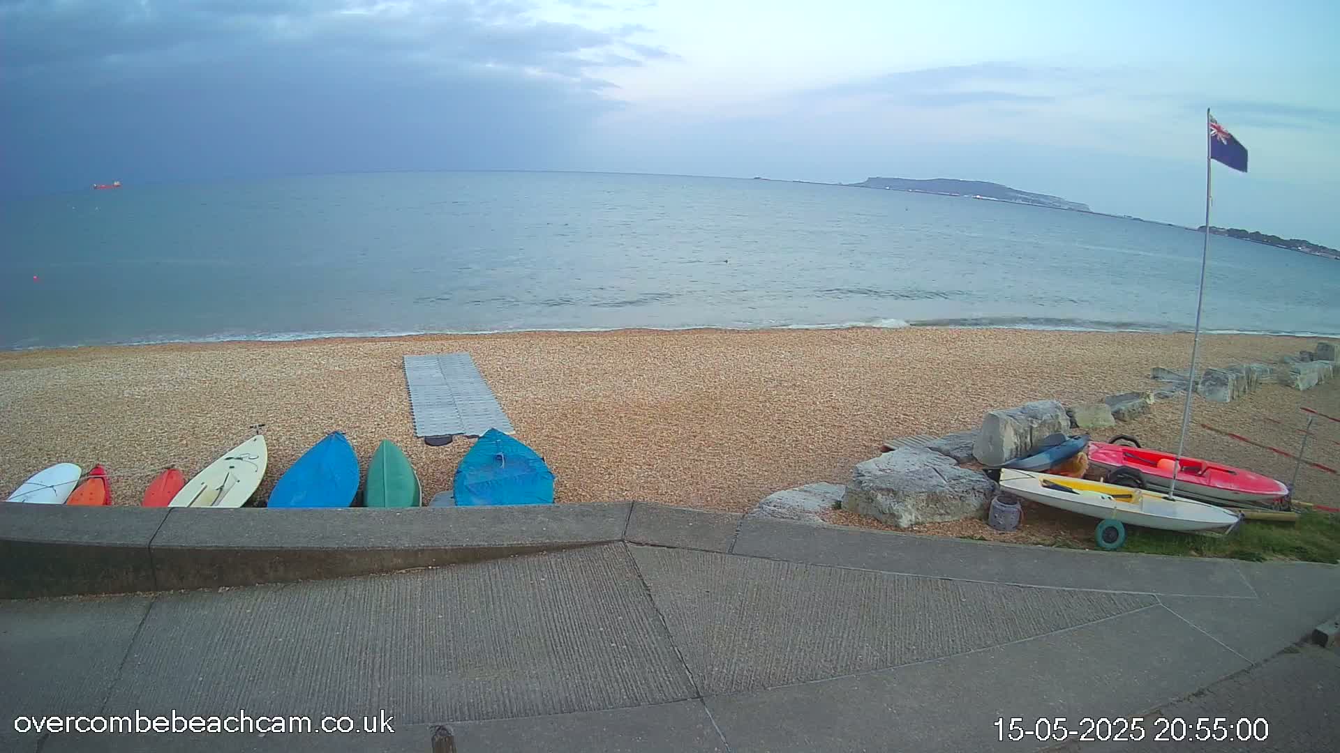 Several small boats are stored on a pebble beach near a calm sea under a cloudy sky.