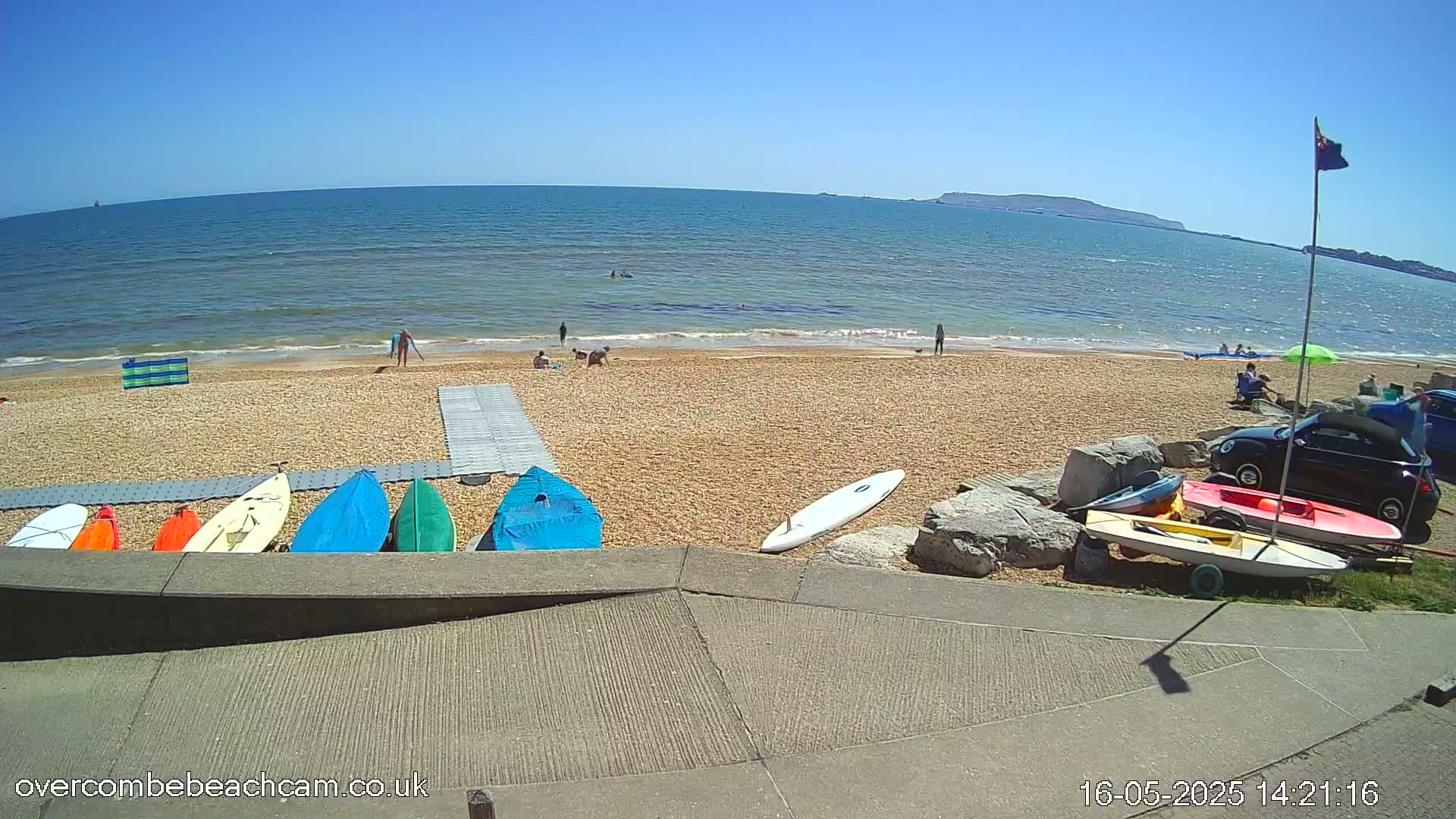 A sandy beach with several kayaks and paddleboards lined up near a walkway, overlooking a calm ocean under a clear blue sky, with a few people scattered on the beach and in the water.
