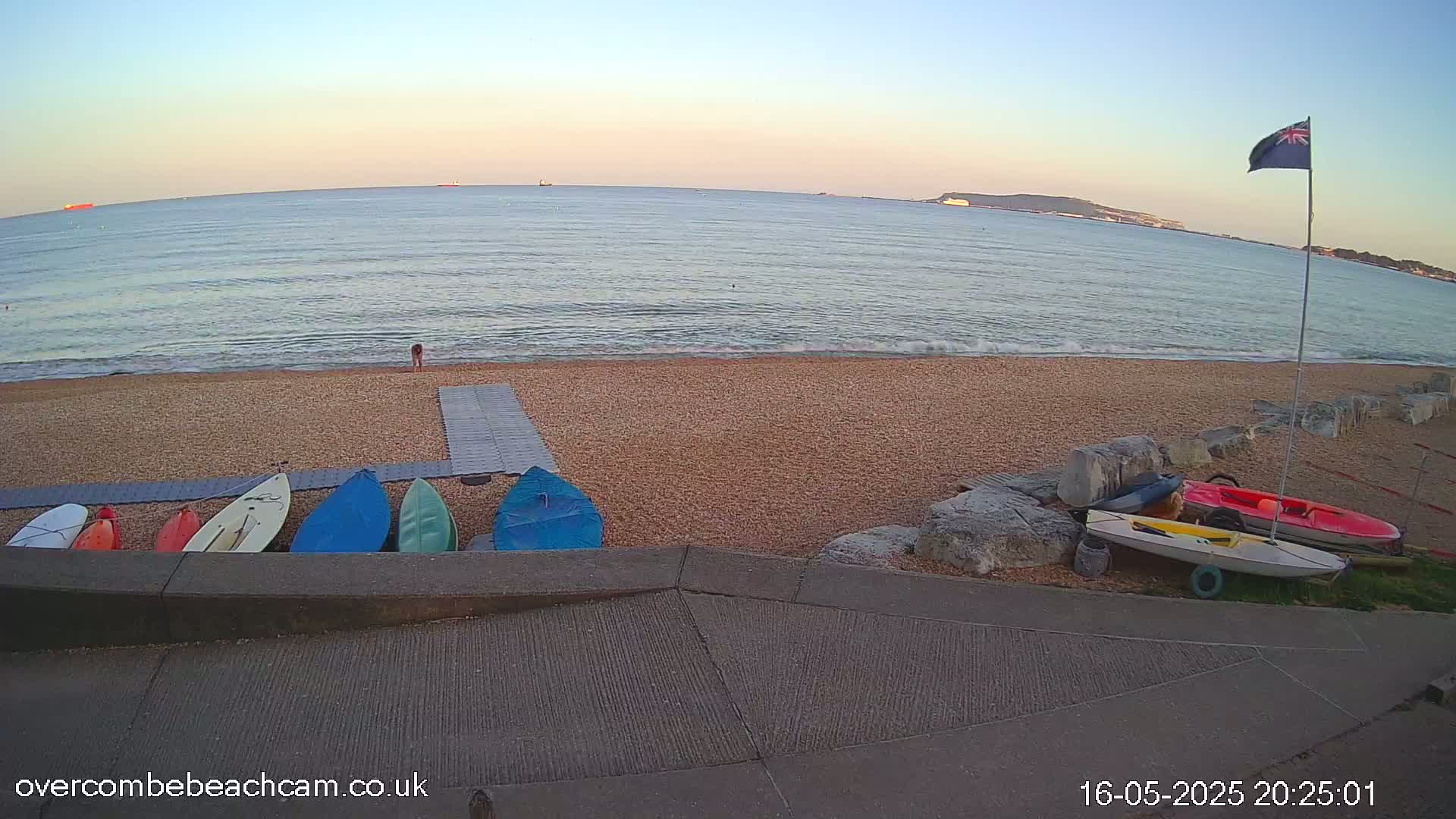 A calm sea under a twilight sky meets a pebbled beach where several kayaks and paddleboards are stored near a ramp, with a single person visible on the sand.