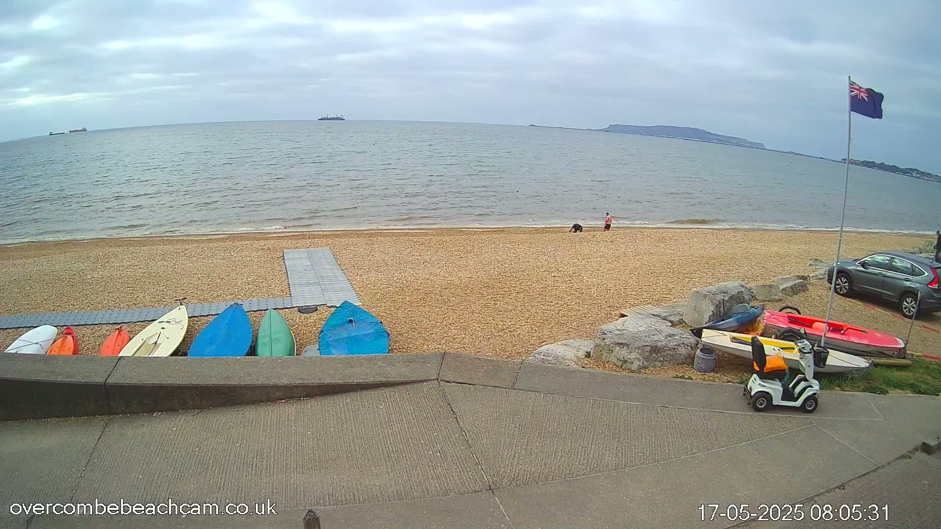 A pebble beach with several kayaks and a mobility scooter, overlooking a calm sea under a cloudy sky.