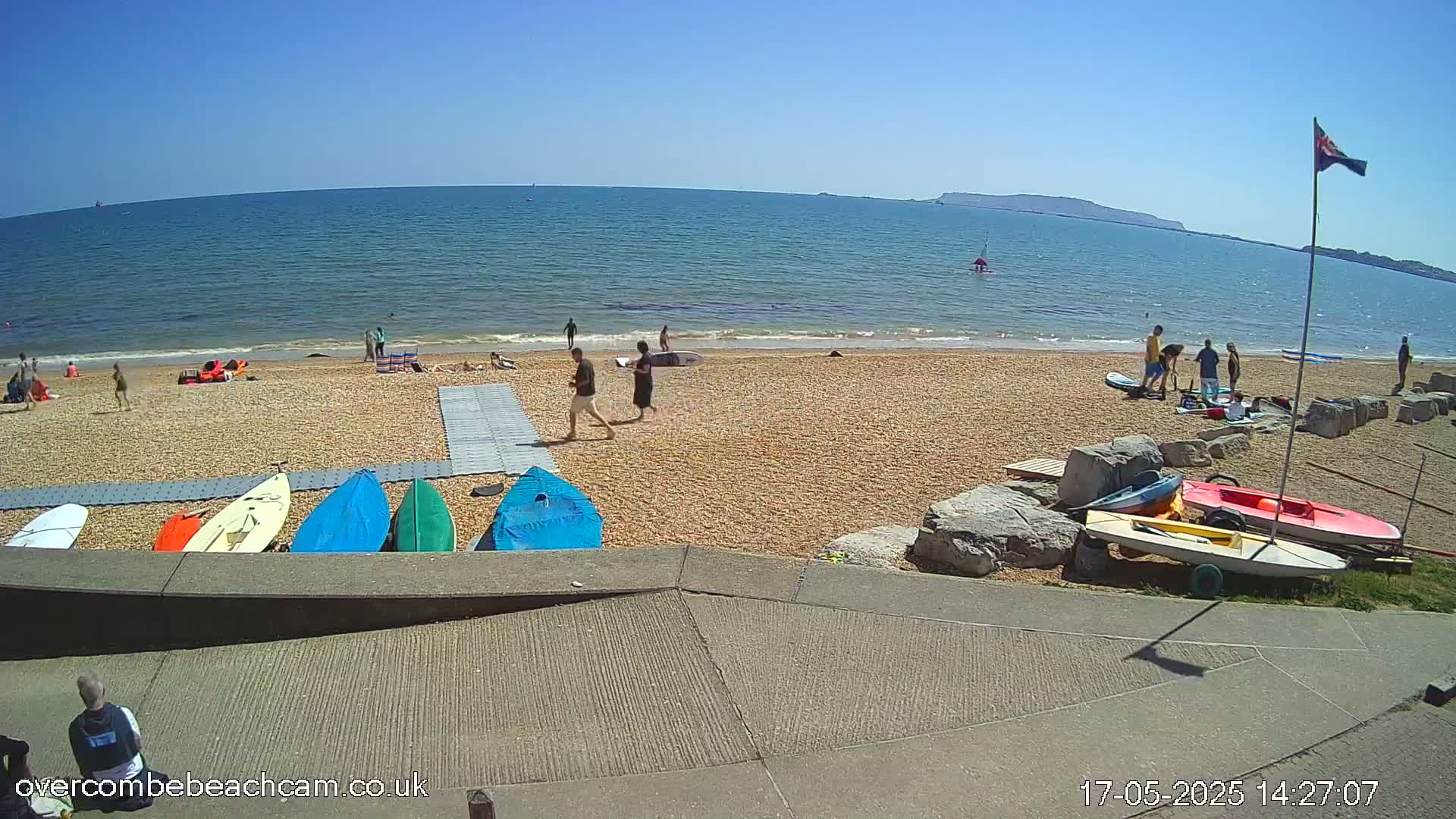 A sunny beach scene shows people relaxing and several small boats and paddleboards on the sand near a calm ocean under a clear blue sky.