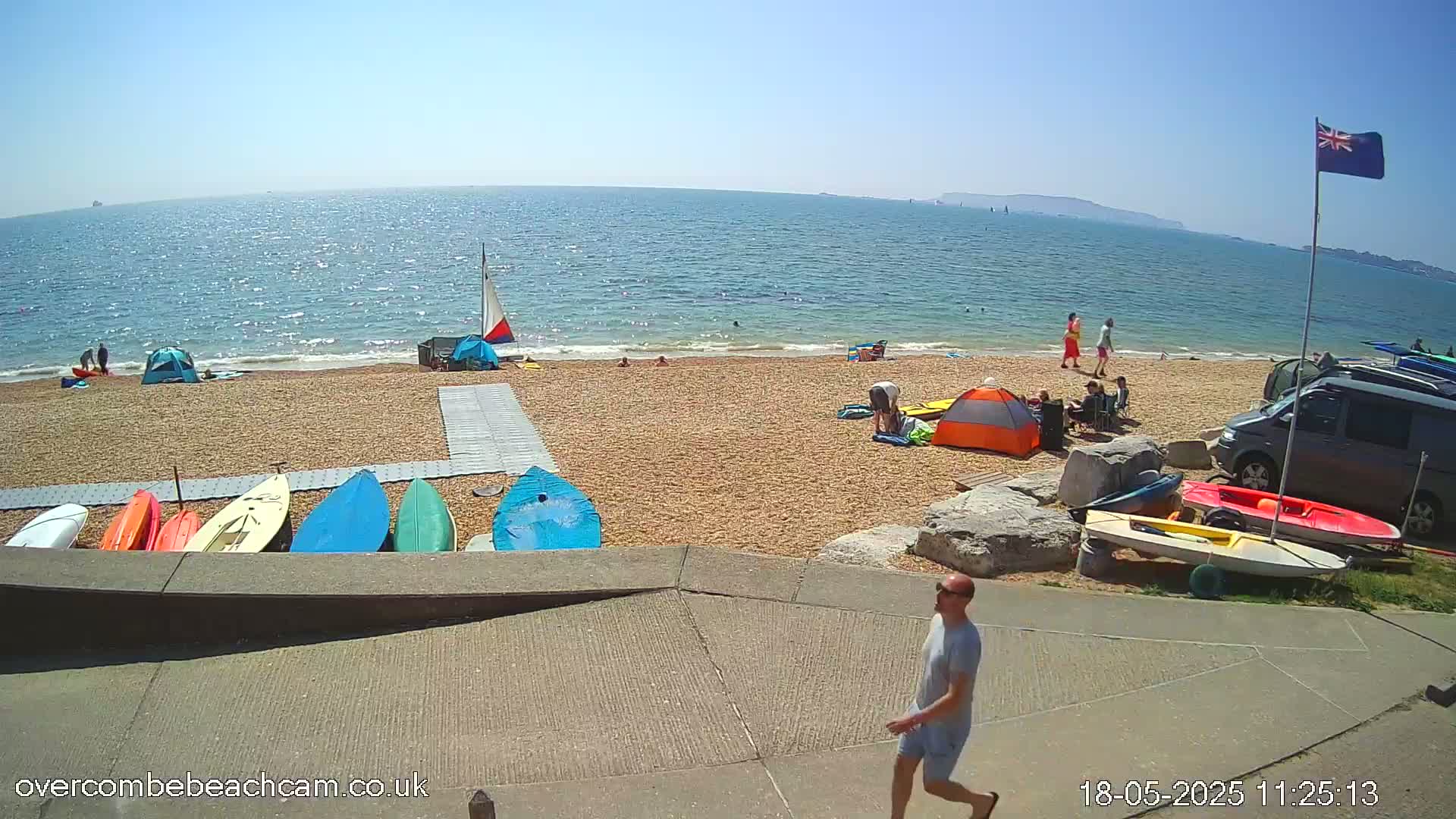 A sunny beach scene shows people relaxing, swimming, and setting up tents near parked cars and a variety of kayaks and sailboats on the shore.
