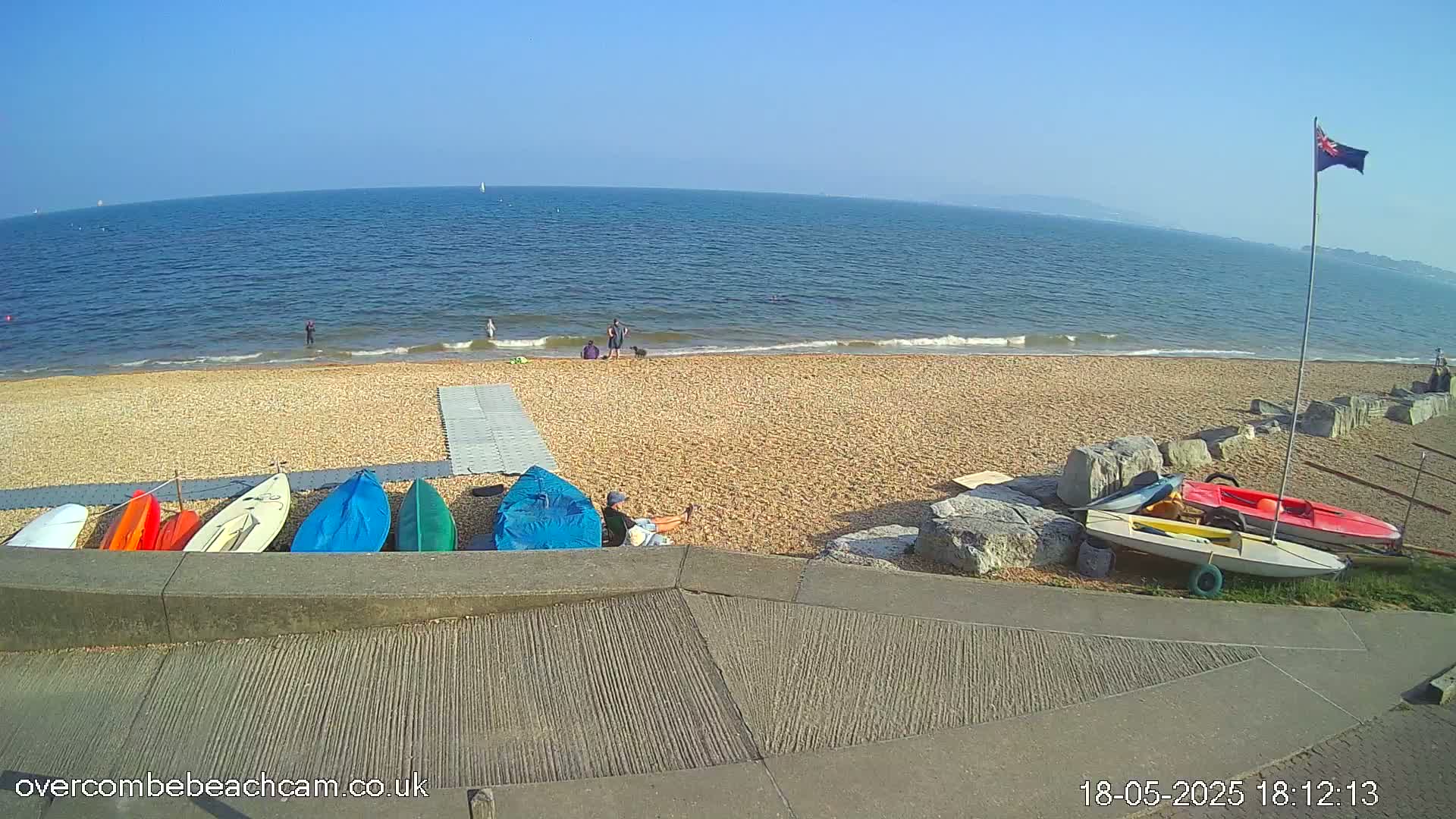 A sandy beach with several small boats on the shore, a few people relaxing and a calm sea under a clear blue sky.