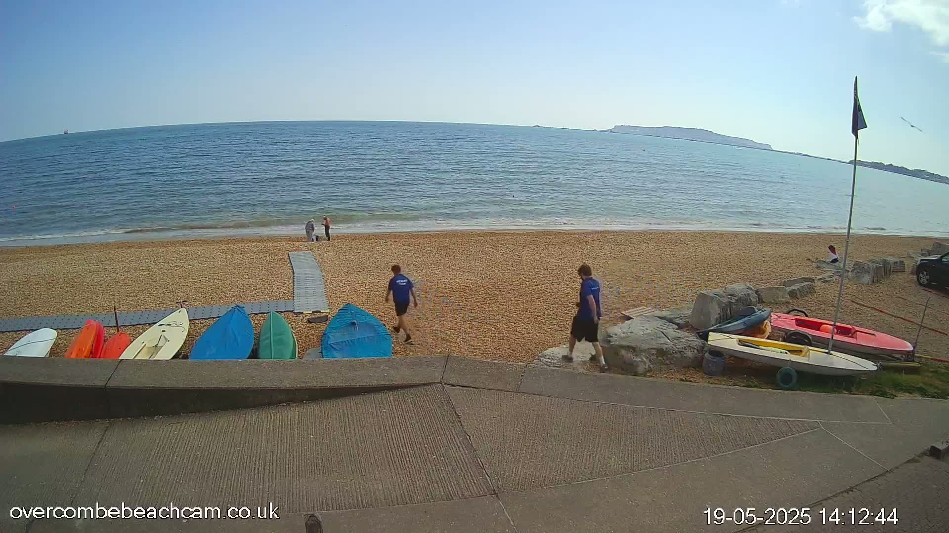 A sandy beach with a calm sea under a clear blue sky, several small boats are lined up on the shore near two people walking away from the water.