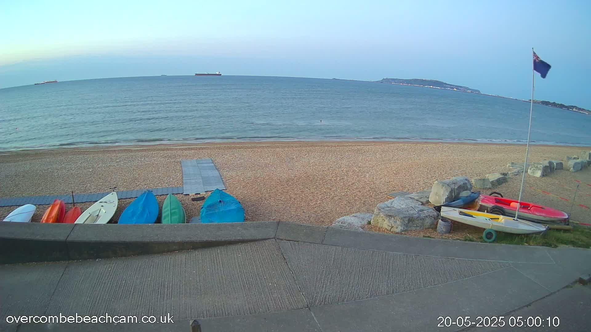 Several small boats are lined up on a pebble beach next to calm ocean water under a clear sky.