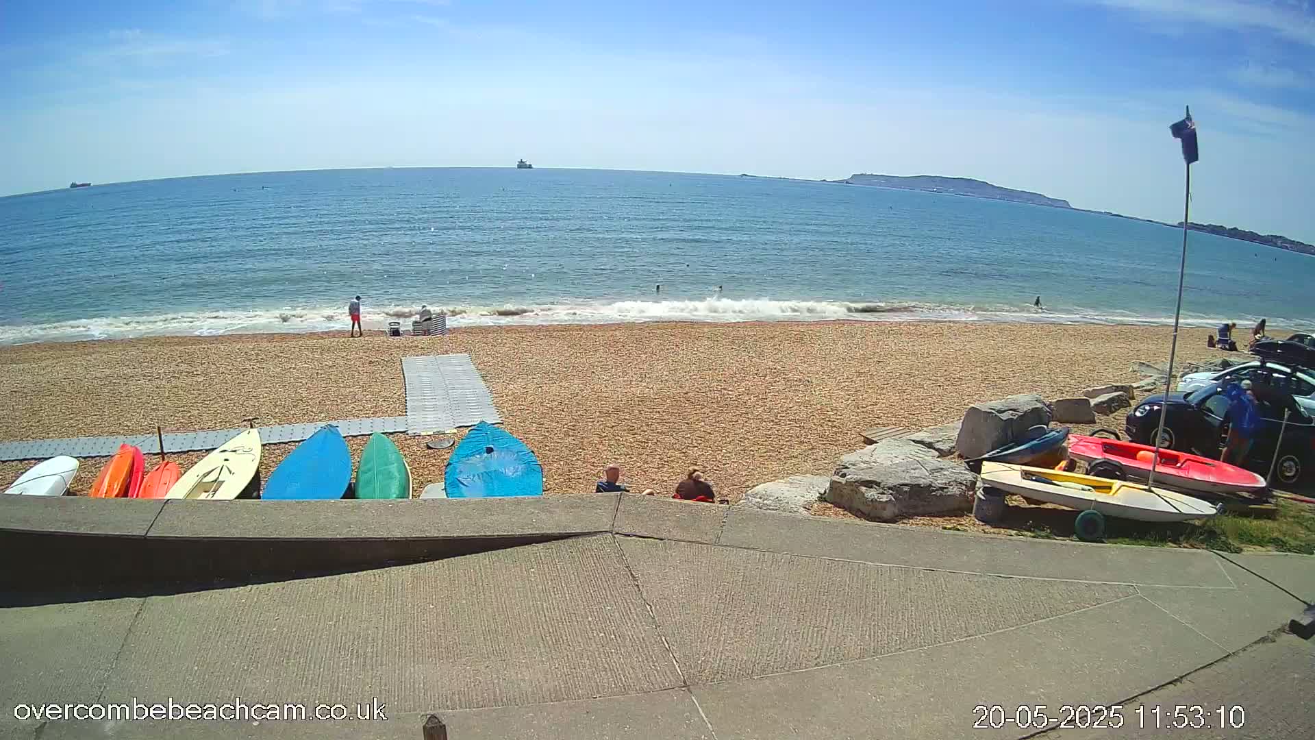 A sunny day at a pebble beach shows several small boats and kayaks on the shore near some people, with a calm sea and distant land visible.