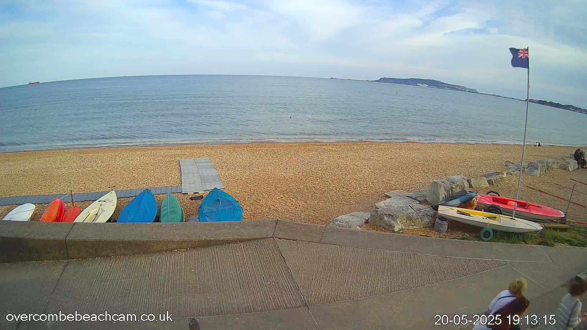 A sandy beach with several small boats and kayaks on the shore, under a partly cloudy sky, overlooks a calm sea with a distant landmass visible on the horizon.