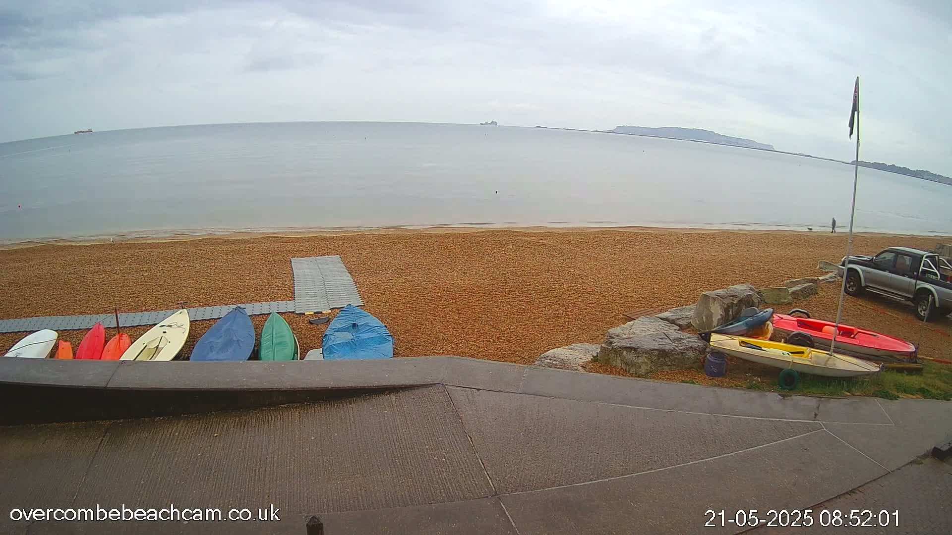 Several small boats and kayaks are lined up on a pebble beach near calm, gray water under an overcast sky.