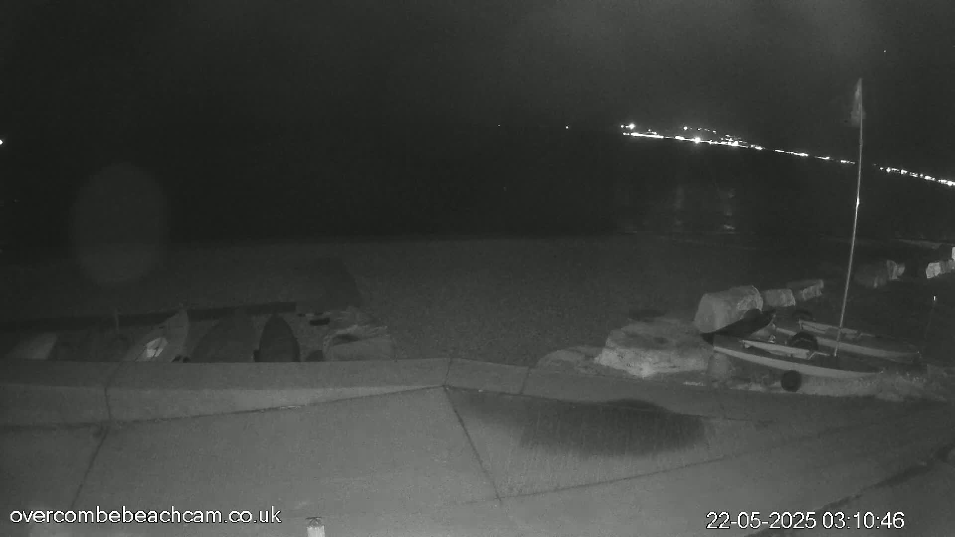 A nighttime, low-light view of a shoreline shows several small boats resting near a low wall, with faint lights visible in the distance across the water.