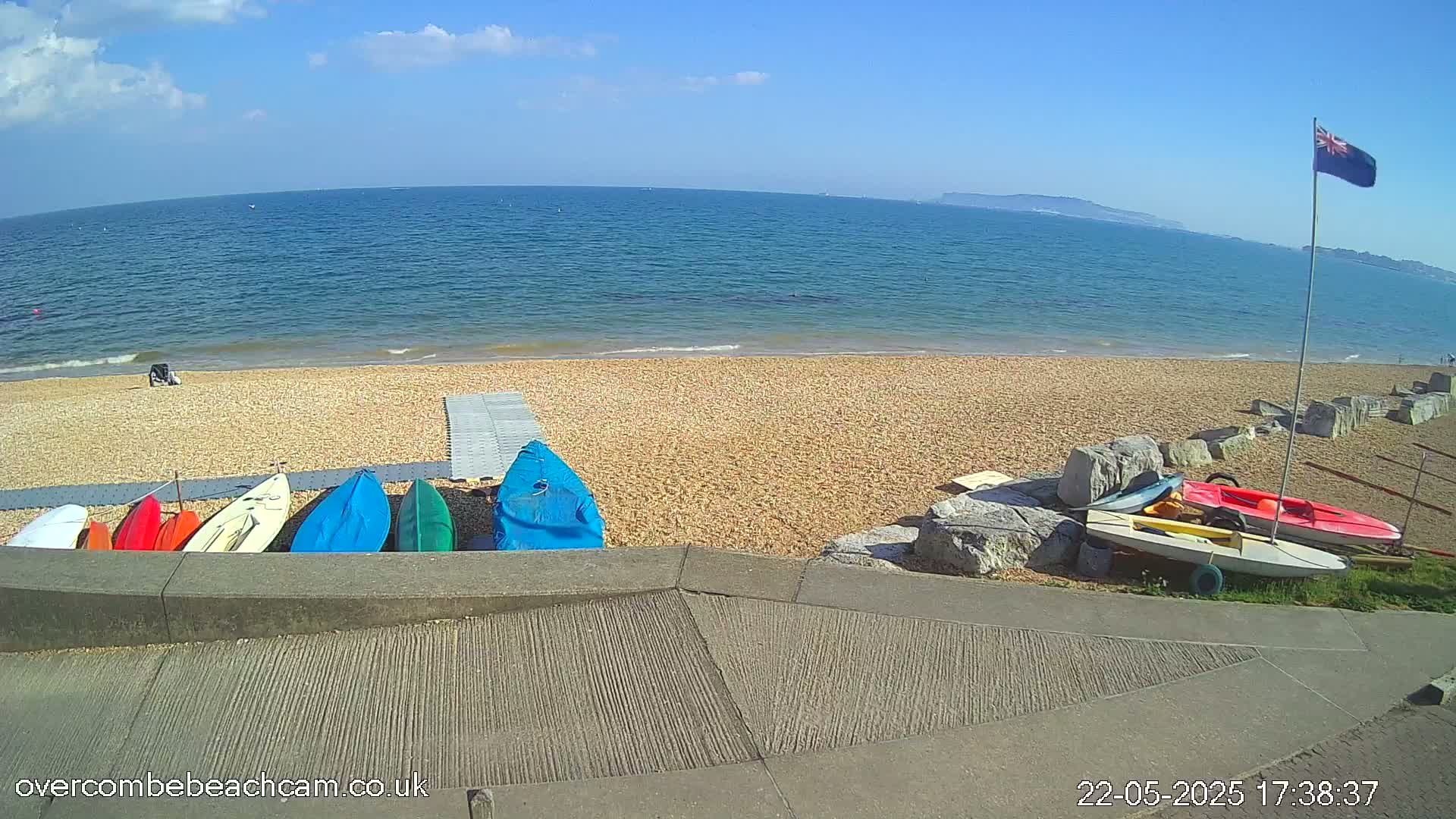 Several colorful kayaks and a small sailboat are stored on a pebble beach next to a calm blue ocean under a sunny sky.