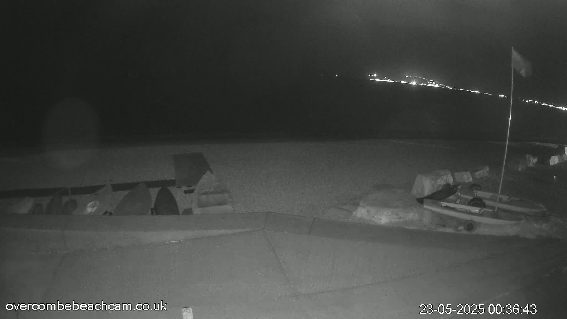 A nighttime, low-light view of a beach showing several upturned boats near a low wall, a small boat-like object on the sand, and distant lights across the water.