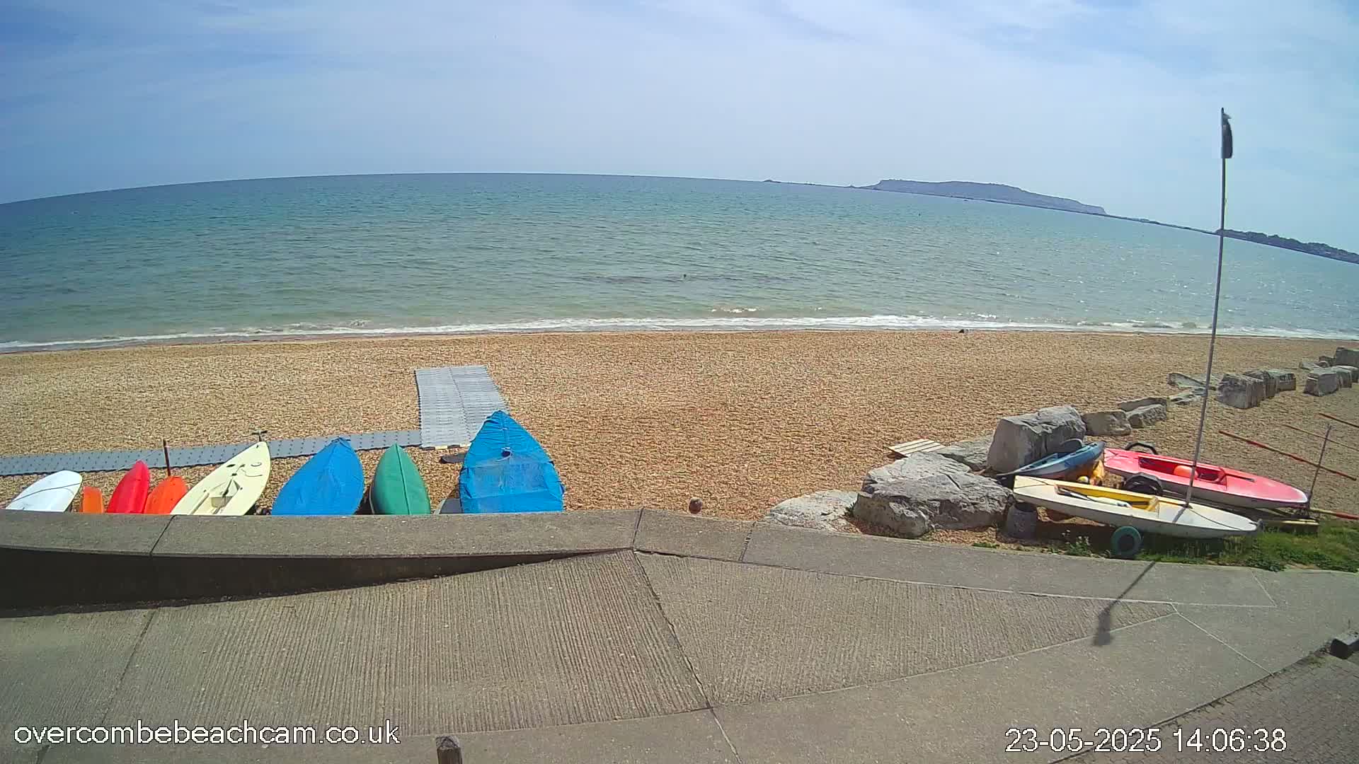 Several small boats and kayaks are lined up on a sandy beach next to calm, blue ocean under a sunny sky.