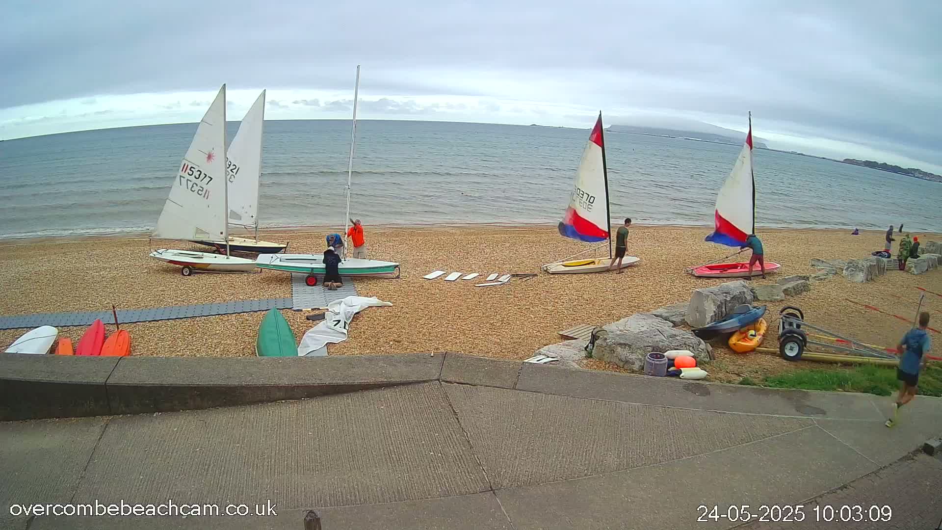Several sailboats are beached on a pebble shoreline under an overcast sky, with people near them and kayaks nearby.