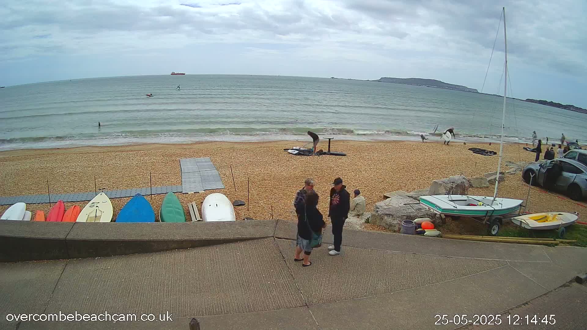 A pebble beach with several small boats and kayaks on the shore, a sailboat, and several people standing near a walkway, all under an overcast sky.