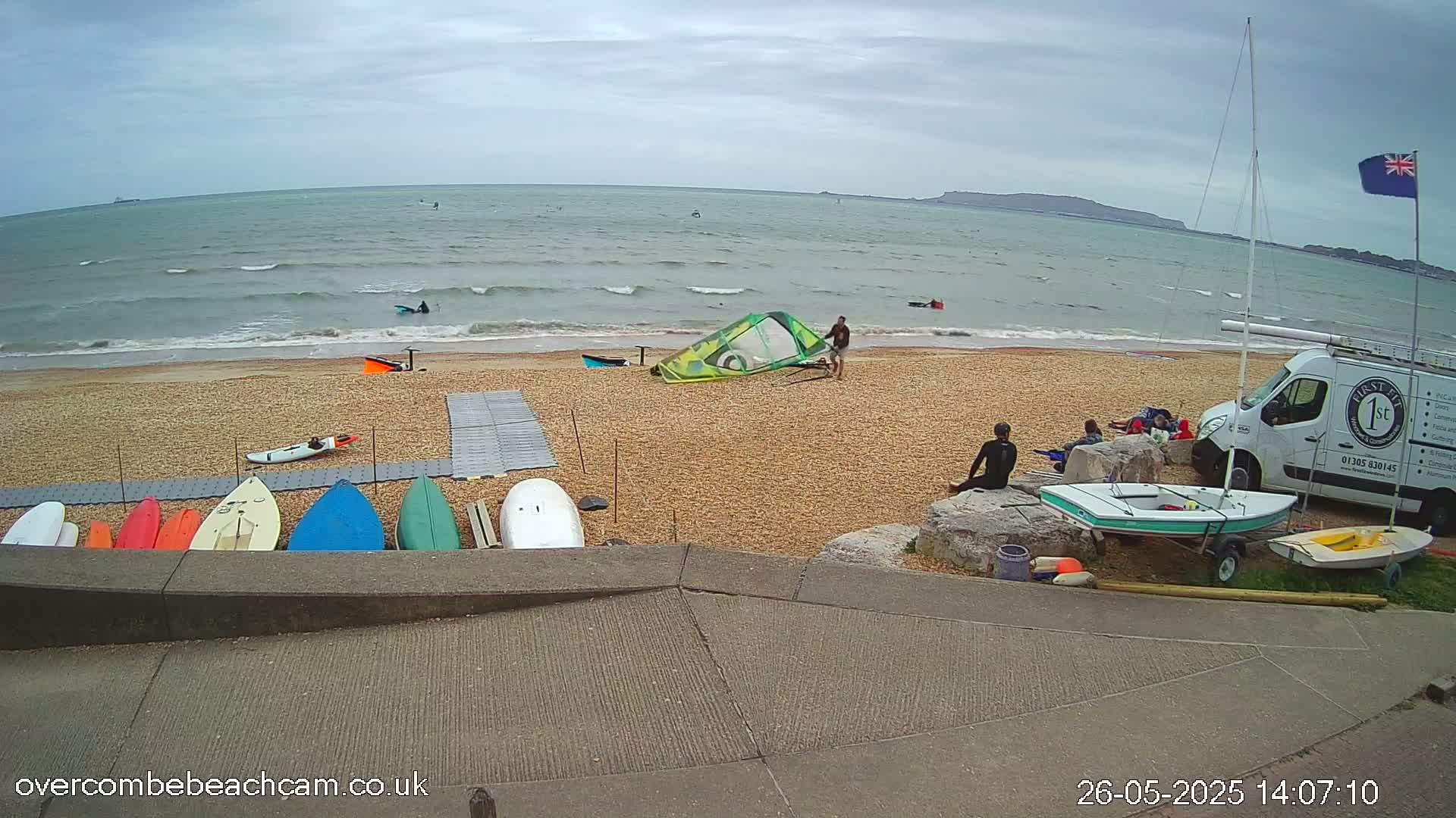 A pebble beach with several people, various water sports equipment including sailboards and kayaks, a van, and a sailboat under an overcast sky with a calm sea.