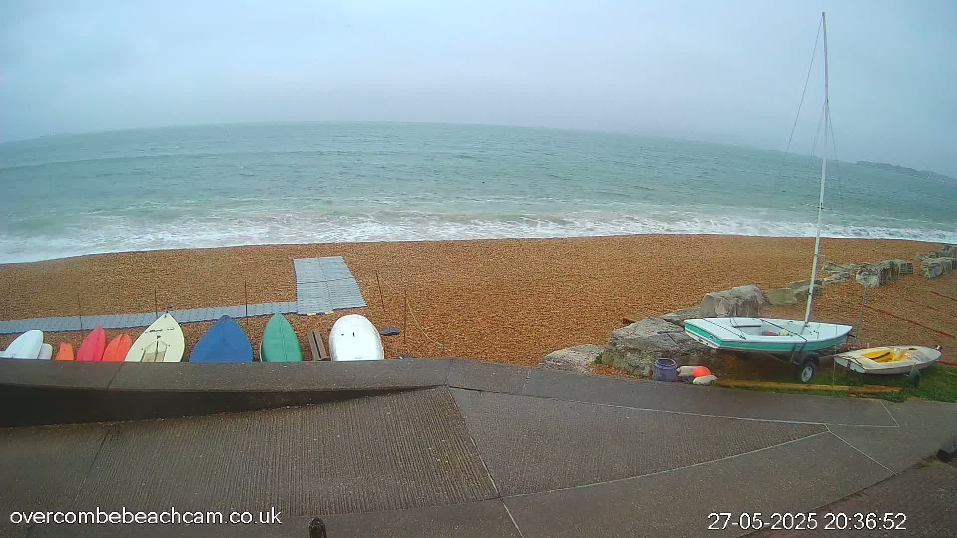 A pebble beach with a small sailboat and several kayaks and paddleboards on the shore, under an overcast sky with a slightly wavy sea.