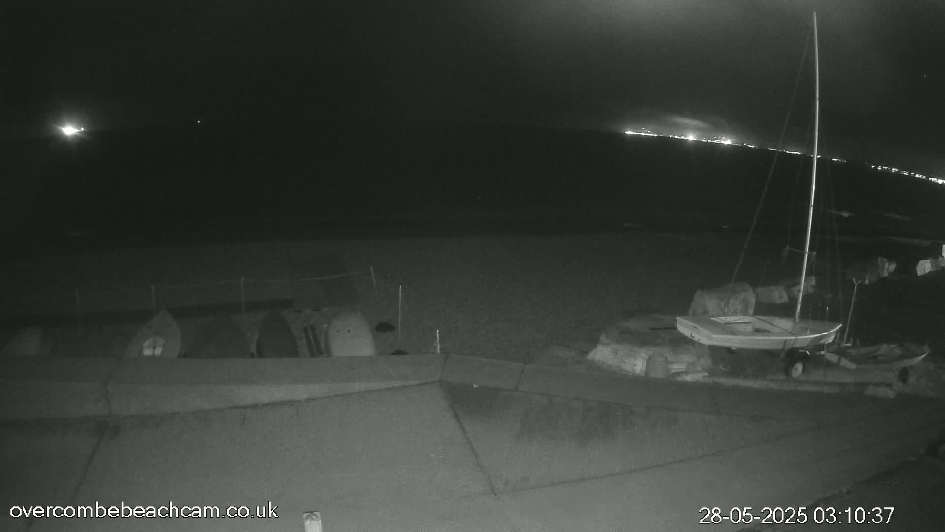 A nighttime, low-light image shows a sailboat beached on rocks near several objects resembling overturned small boats or shelters on a dark beach under a mostly clear sky with distant lights visible on the horizon.
