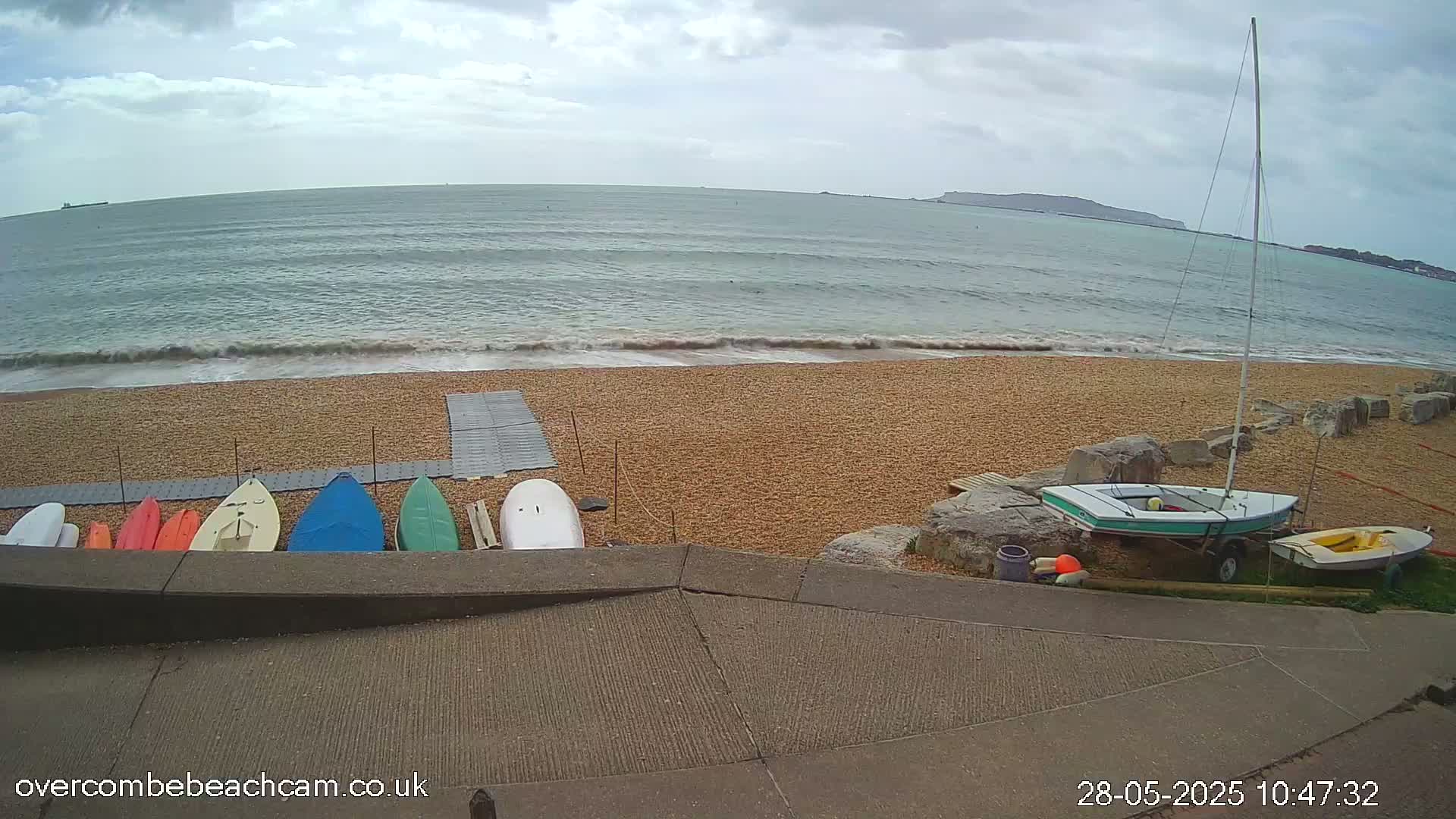 A pebble beach with small waves under a cloudy sky shows several kayaks and paddleboards lined up on the shore, along with a small sailboat on a trailer.