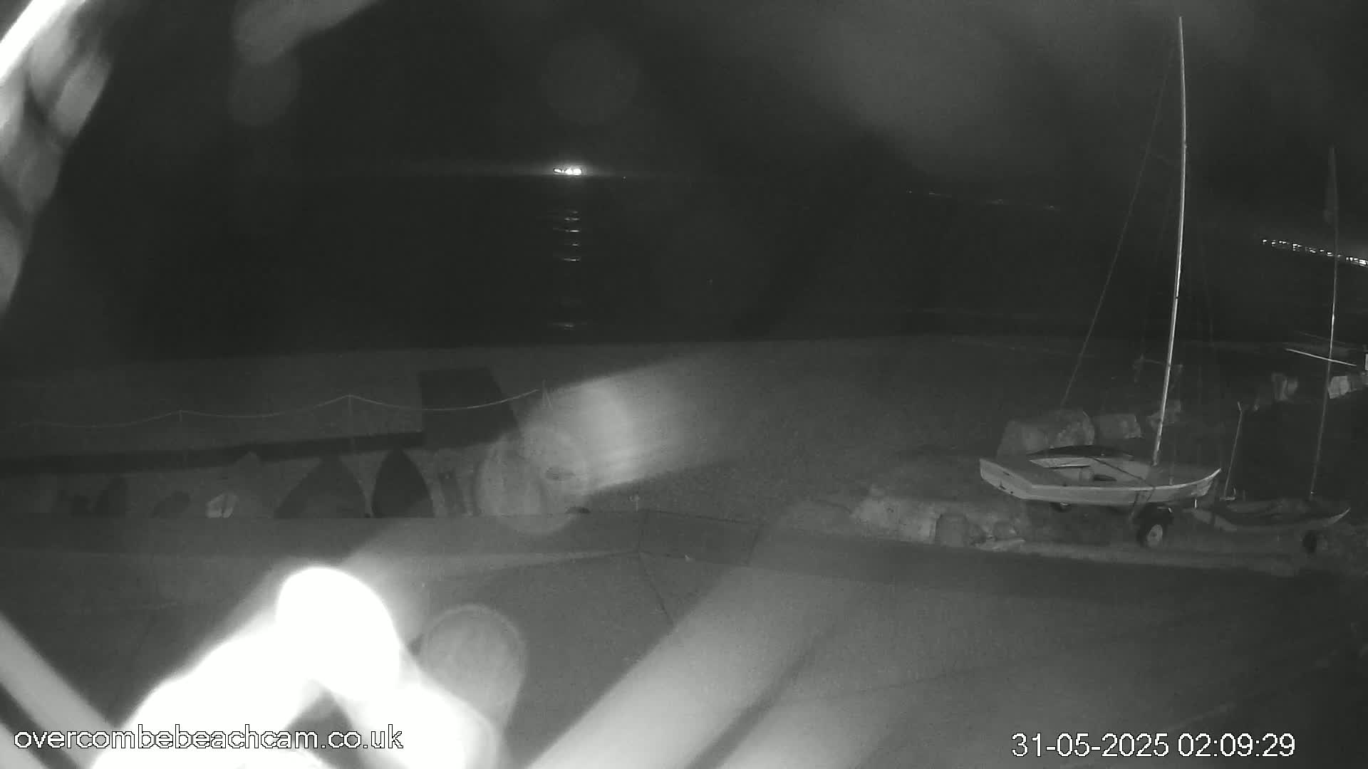 A nighttime, low-light image shows a small sailboat on a trailer near a dark beach with a calm sea in the background.