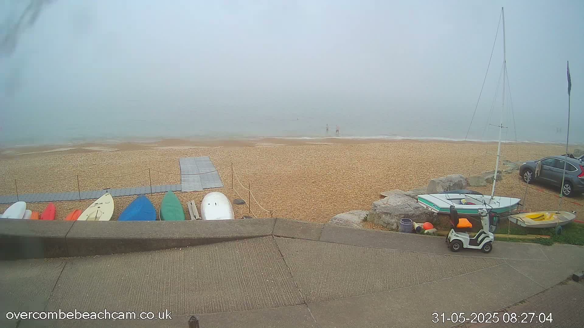 A foggy beach scene shows several small boats and kayaks stored on a pebble beach near a parked car and a mobility scooter.