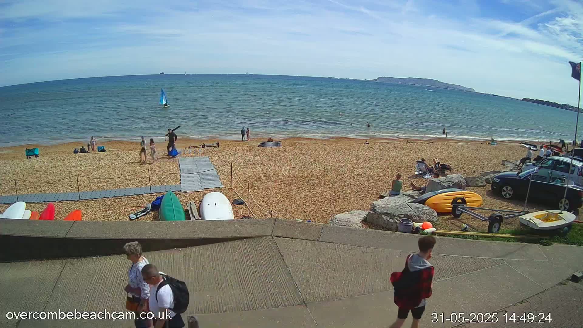 A sunny beach scene shows people sunbathing, swimming, and walking near the water, with several kayaks and a sailboat visible, and some people walking away from the beach on a paved path.