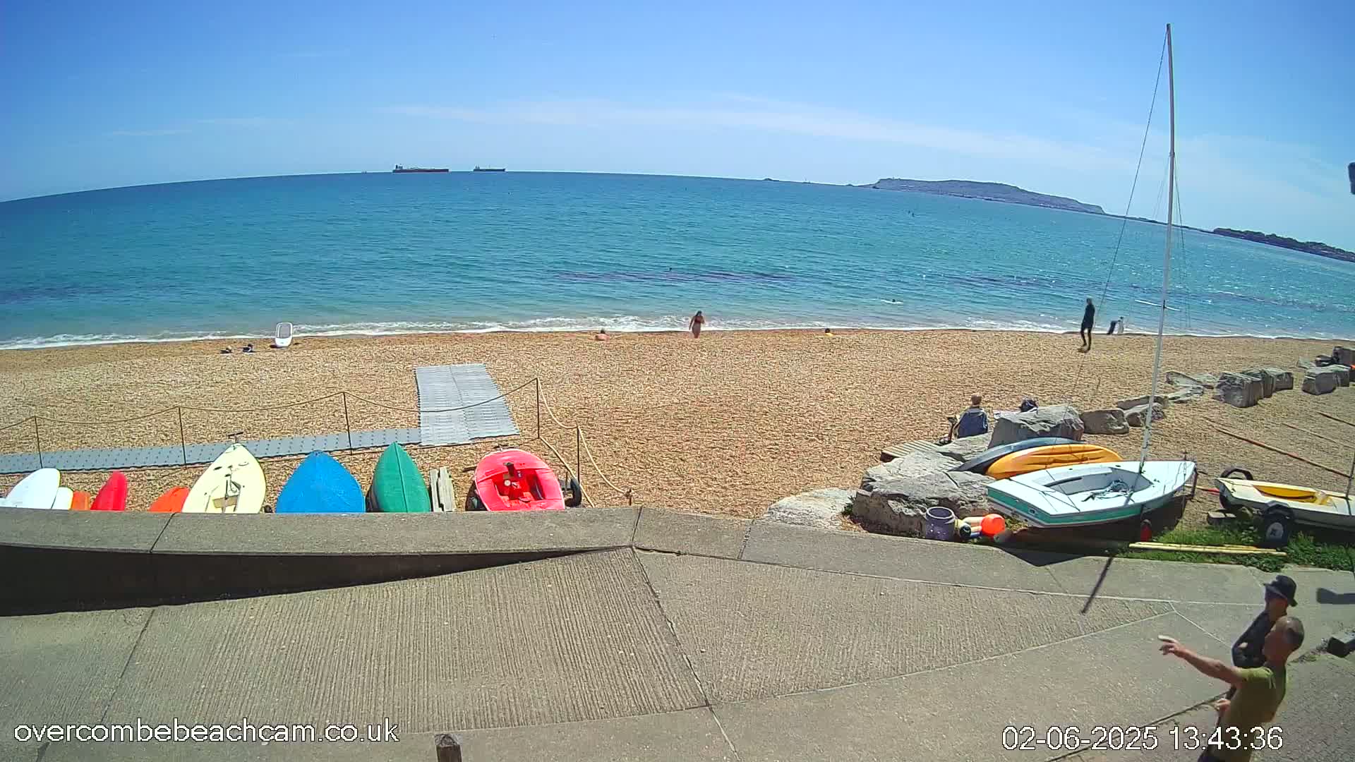 A sandy beach with colorful kayaks and a sailboat on a sunny day, overlooking a calm ocean with several people visible.