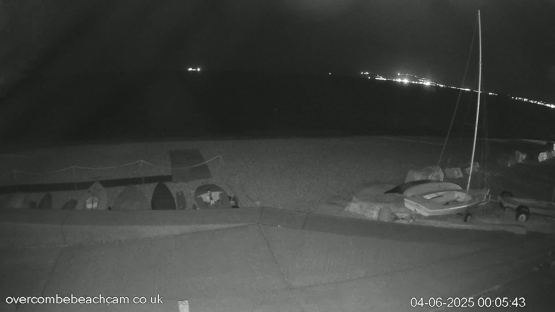 A nighttime, low-light view of a beach shows several boats and other watercraft stored near a seawall, with distant lights visible across the water.