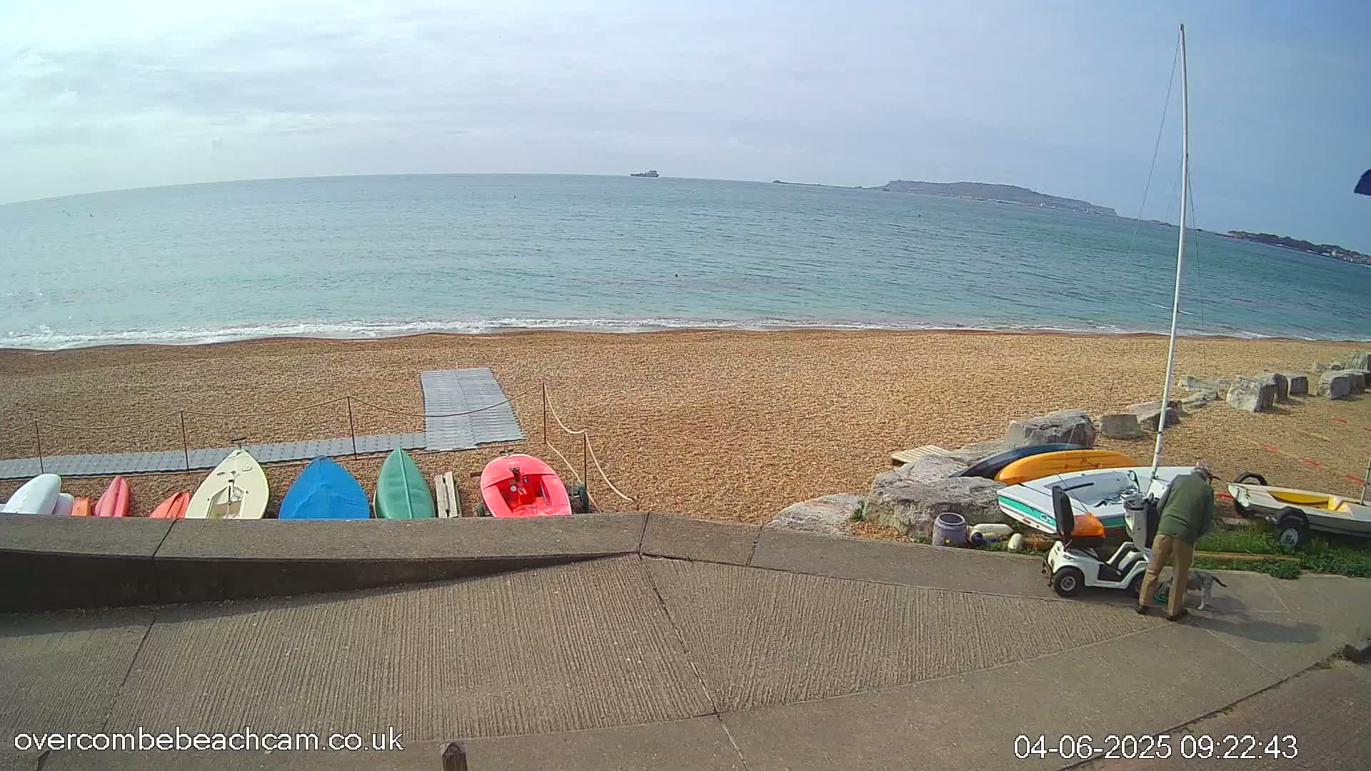 A sandy beach with colorful small boats and a person with a mobility scooter is visible, overlooking a calm sea under a mostly clear sky.