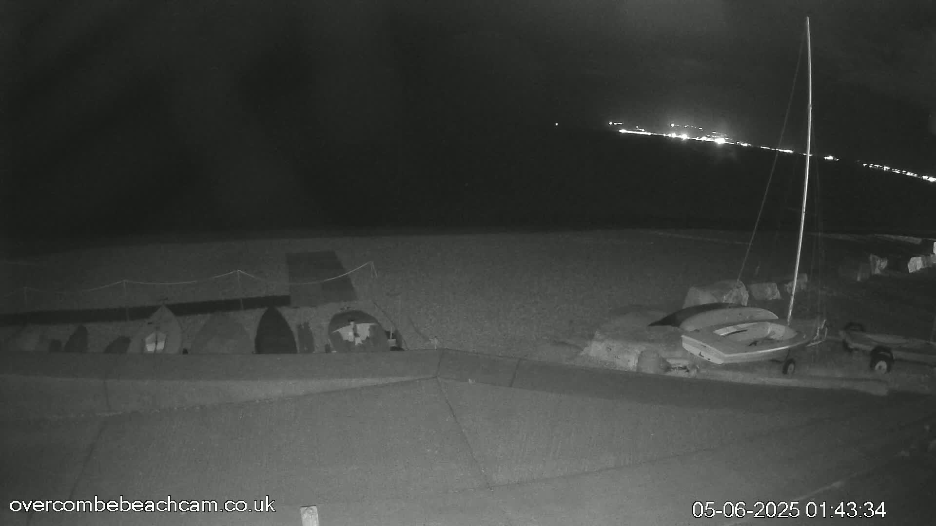 A nighttime, low-light view of a beach shows several small boats beached near a low wall, with a sailboat and a small dinghy nearby, and distant lights visible on the water.