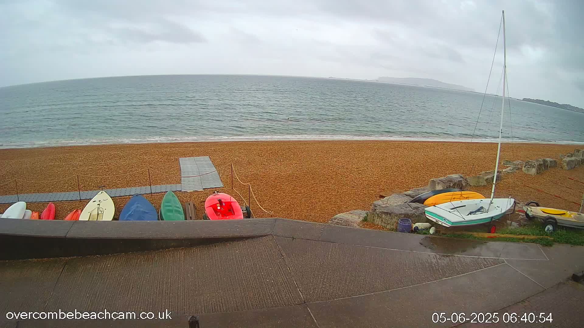A pebble beach with several small boats and kayaks on a wet, overcast day, overlooking a calm sea.