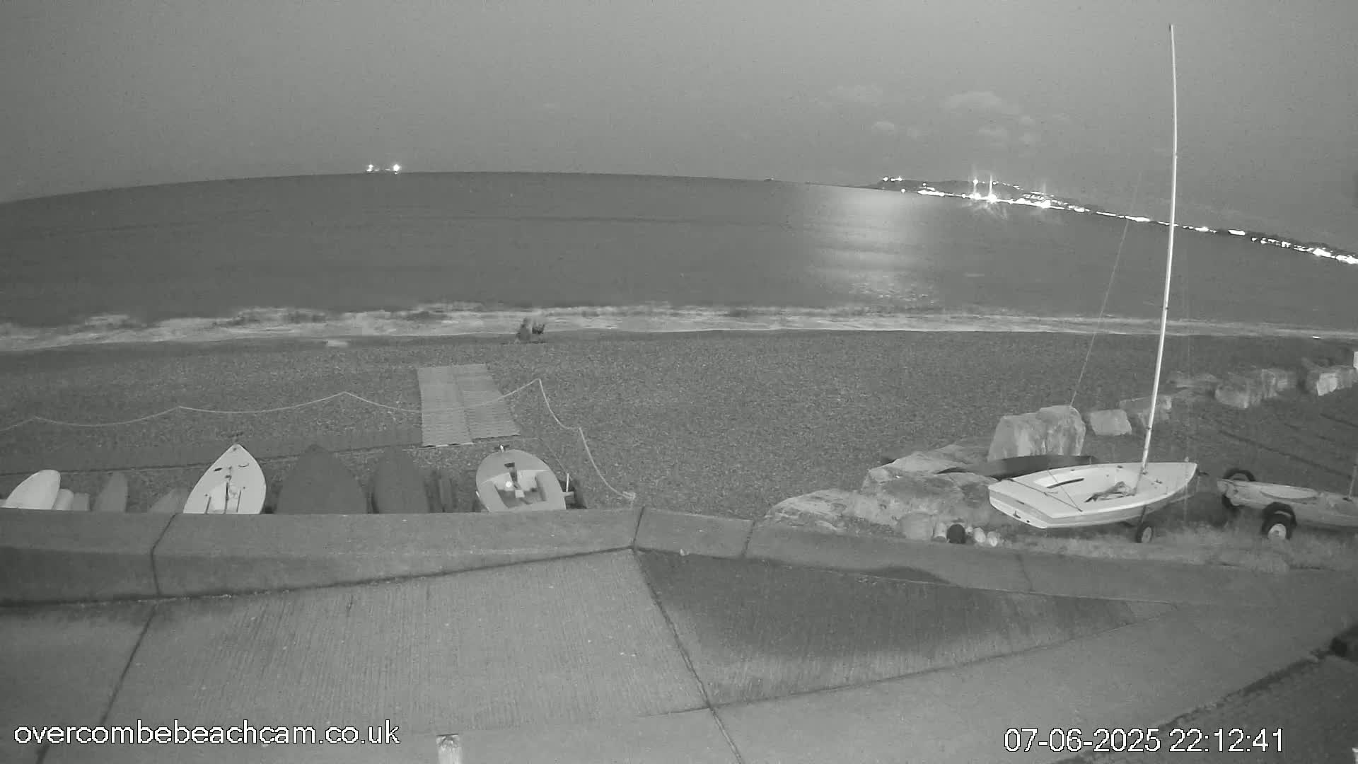 A nighttime, grayscale view of a pebble beach with several small boats on shore, a sailboat, and a calm sea reflecting lights from a distant town under a cloudy sky.