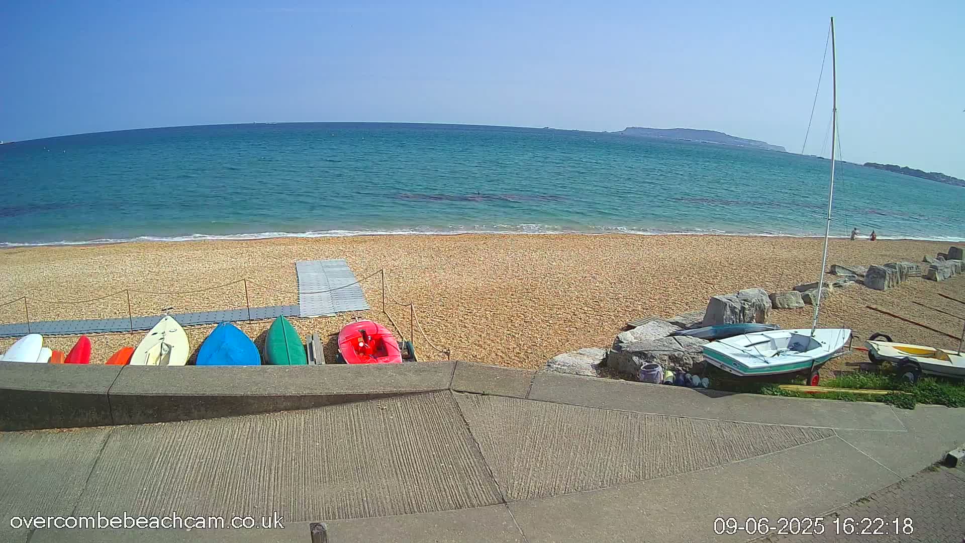 A sandy beach with several small boats and kayaks lined up near a low concrete wall, overlooking a calm, blue ocean under a clear, sunny sky.