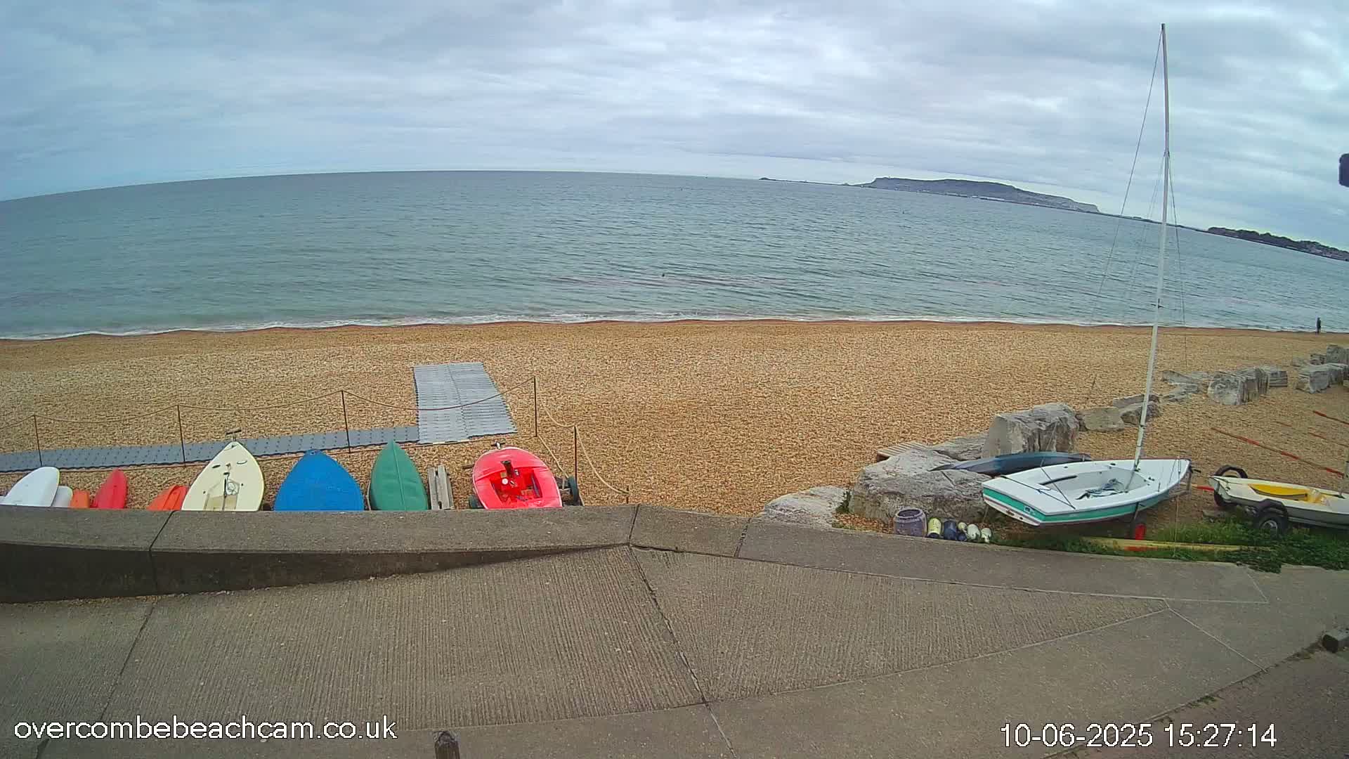 A pebble beach with several small boats and kayaks lined up on the shore, under an overcast sky, overlooks a calm sea with a distant landmass visible.