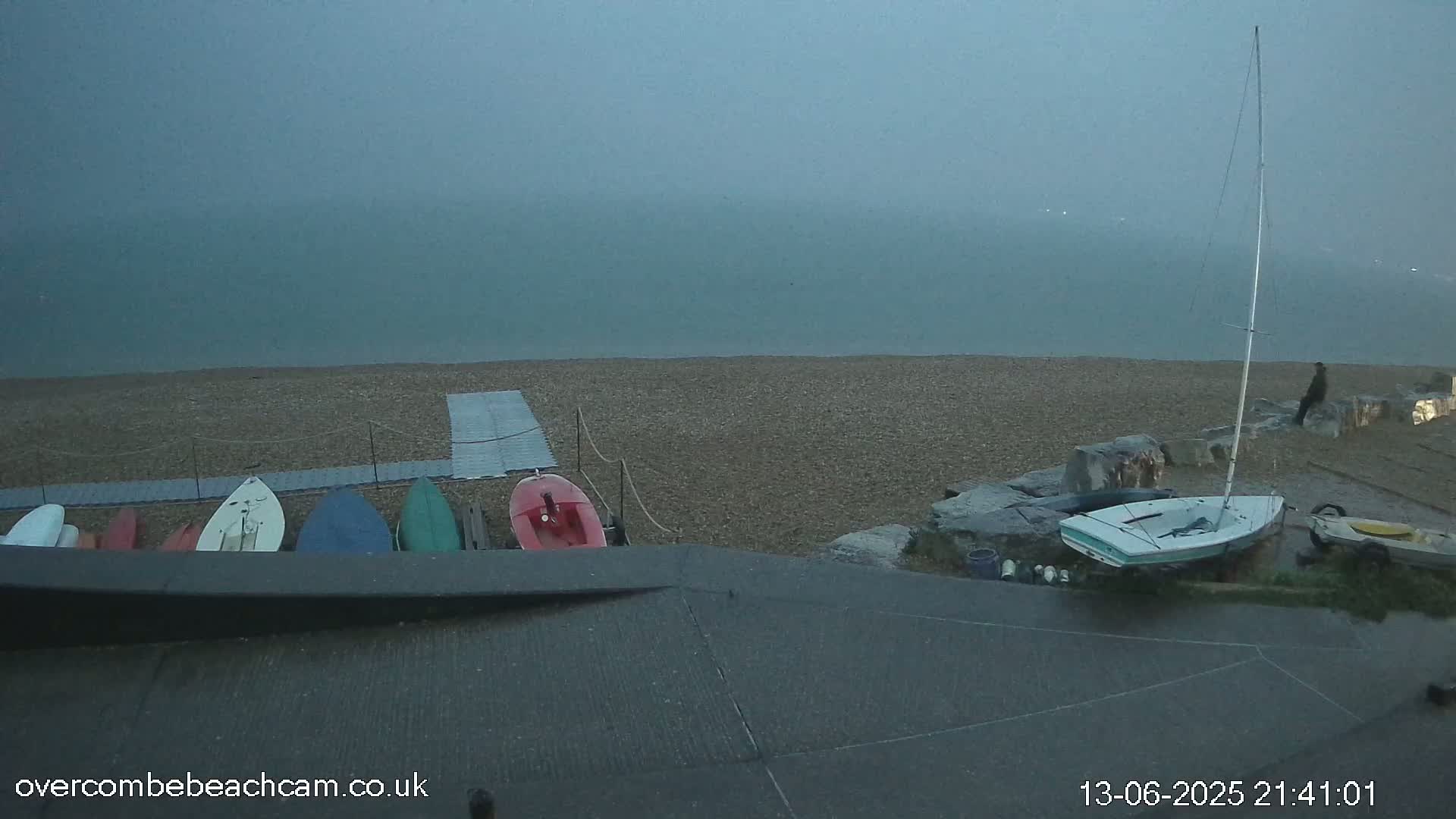 Several small boats are beached on a pebble shore next to a sailboat under a foggy, overcast sky.