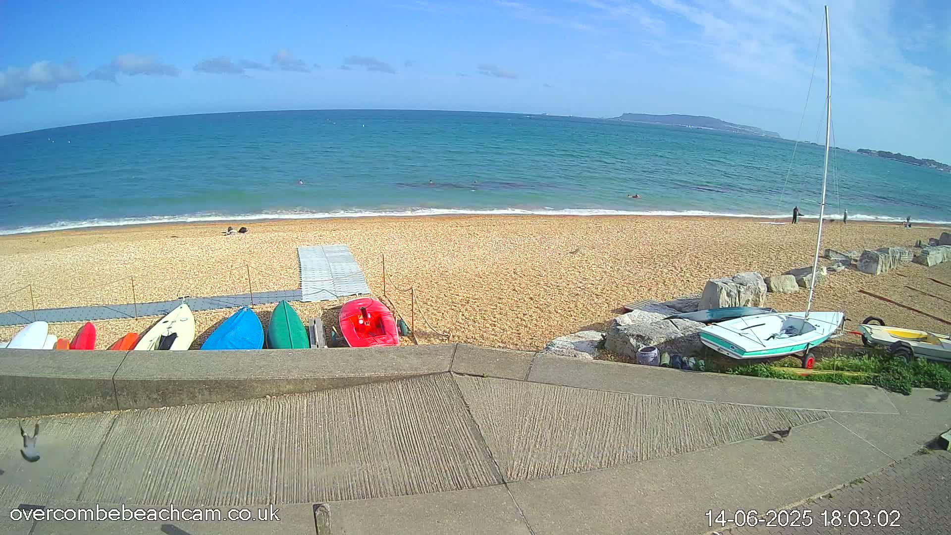 A sandy beach with a calm ocean under a mostly sunny sky, several small boats and kayaks are lined up near a cement ramp.