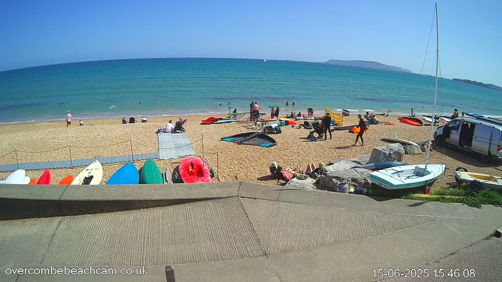 A sunny beach scene shows people relaxing and participating in watersports, with several kayaks and sailboats on the shore.
