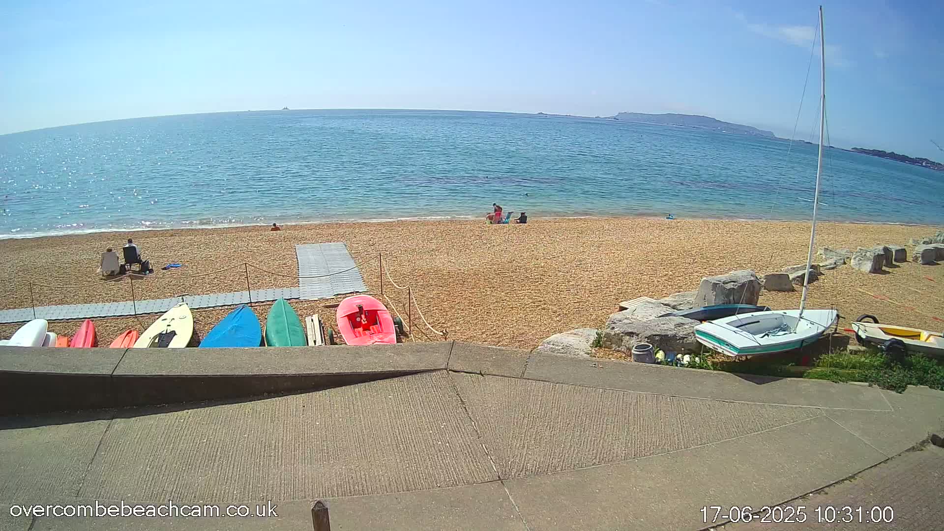 A sandy beach with several small boats and kayaks lined up on the shore, a sailboat beached nearby, and a few people relaxing on the beach under a sunny sky.