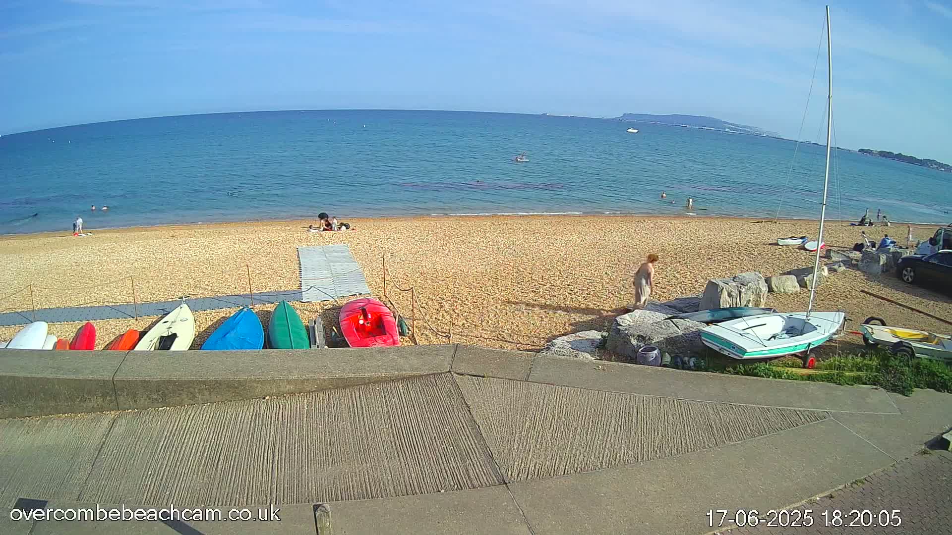 A sandy beach with several kayaks and a sailboat on a sunny day, overlooking a calm ocean with a few people swimming and relaxing.