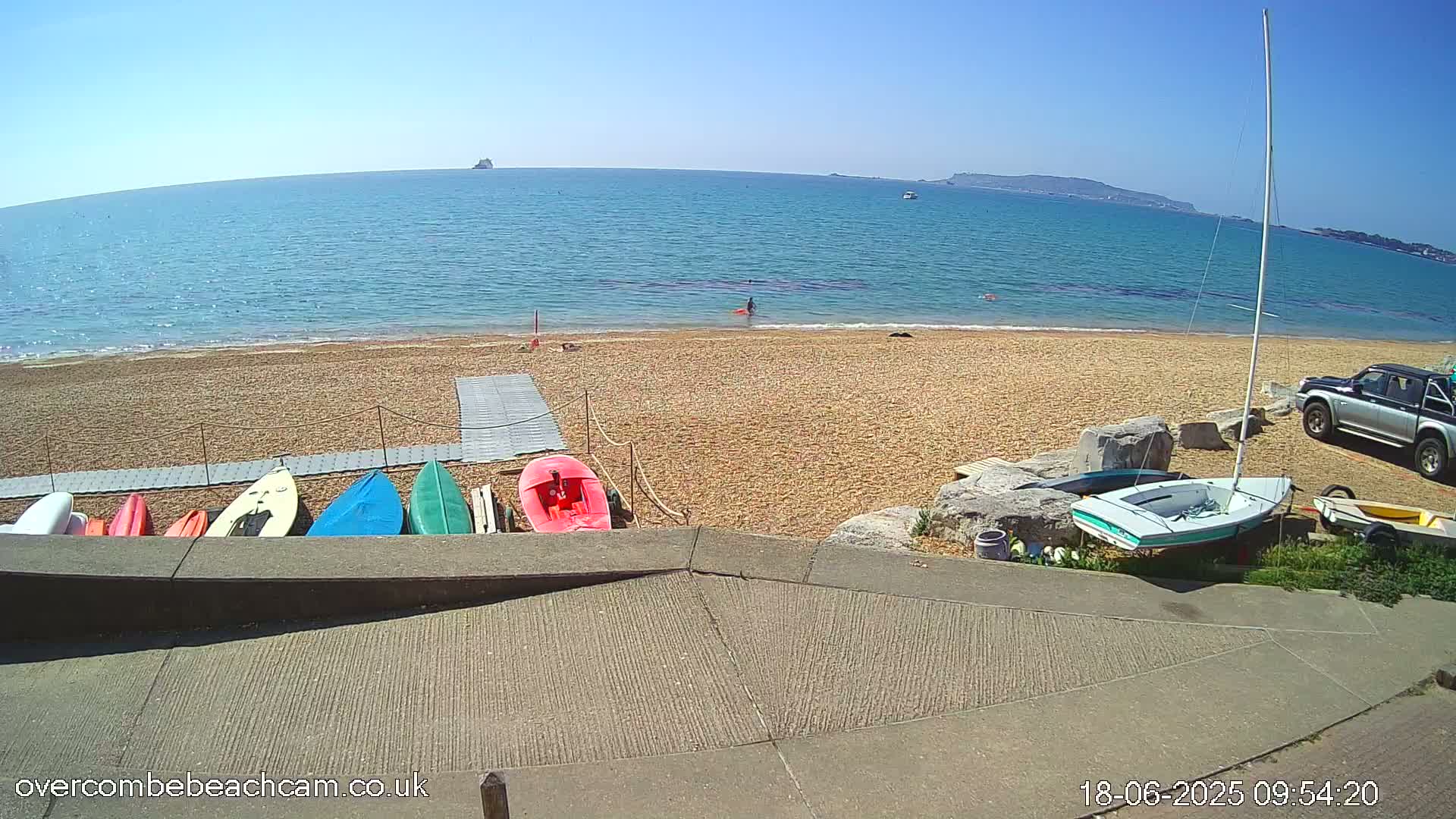 A sunny beach with several small boats and a sailboat on the sand, a person in the water, and a truck parked nearby.
